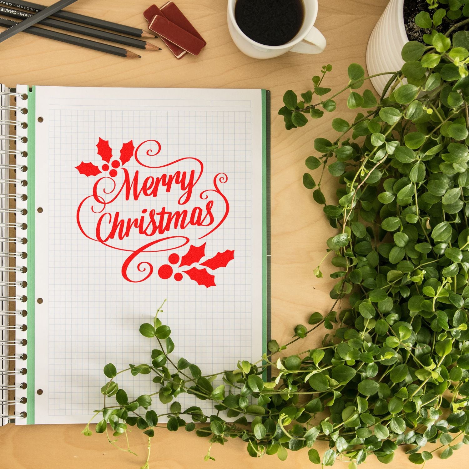 Merry Christmas rubber stamp design in red on graph paper, styled on a desk with coffee, pencils, and greenery.