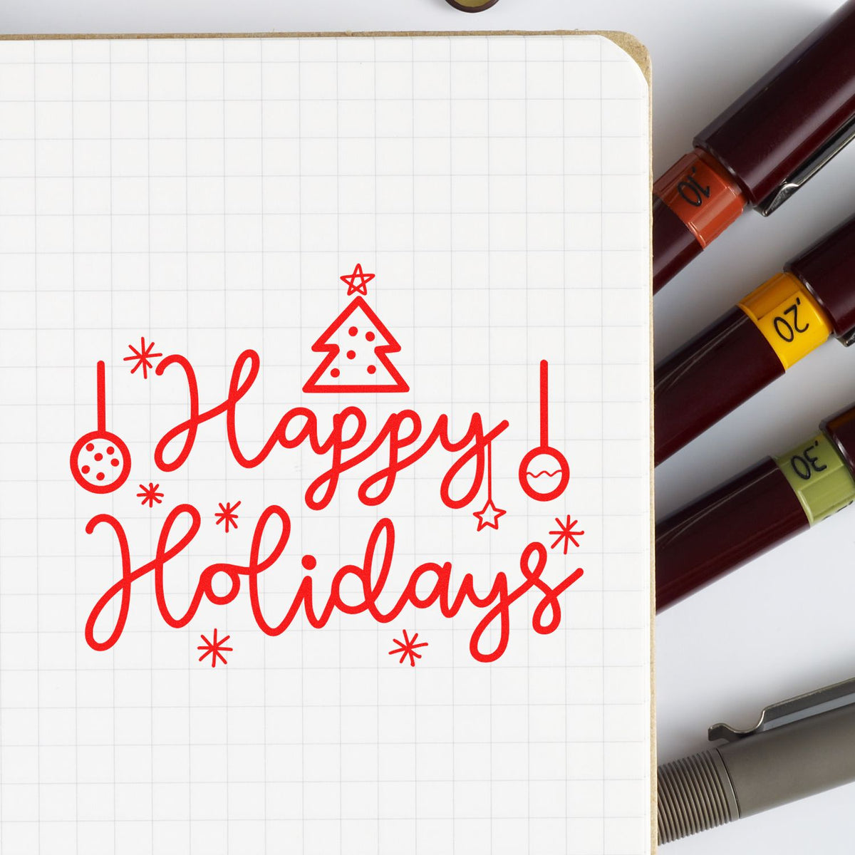 Red Happy Holidays rubber stamp design with Christmas ornaments, shown on a notebook for holiday card making and crafting.