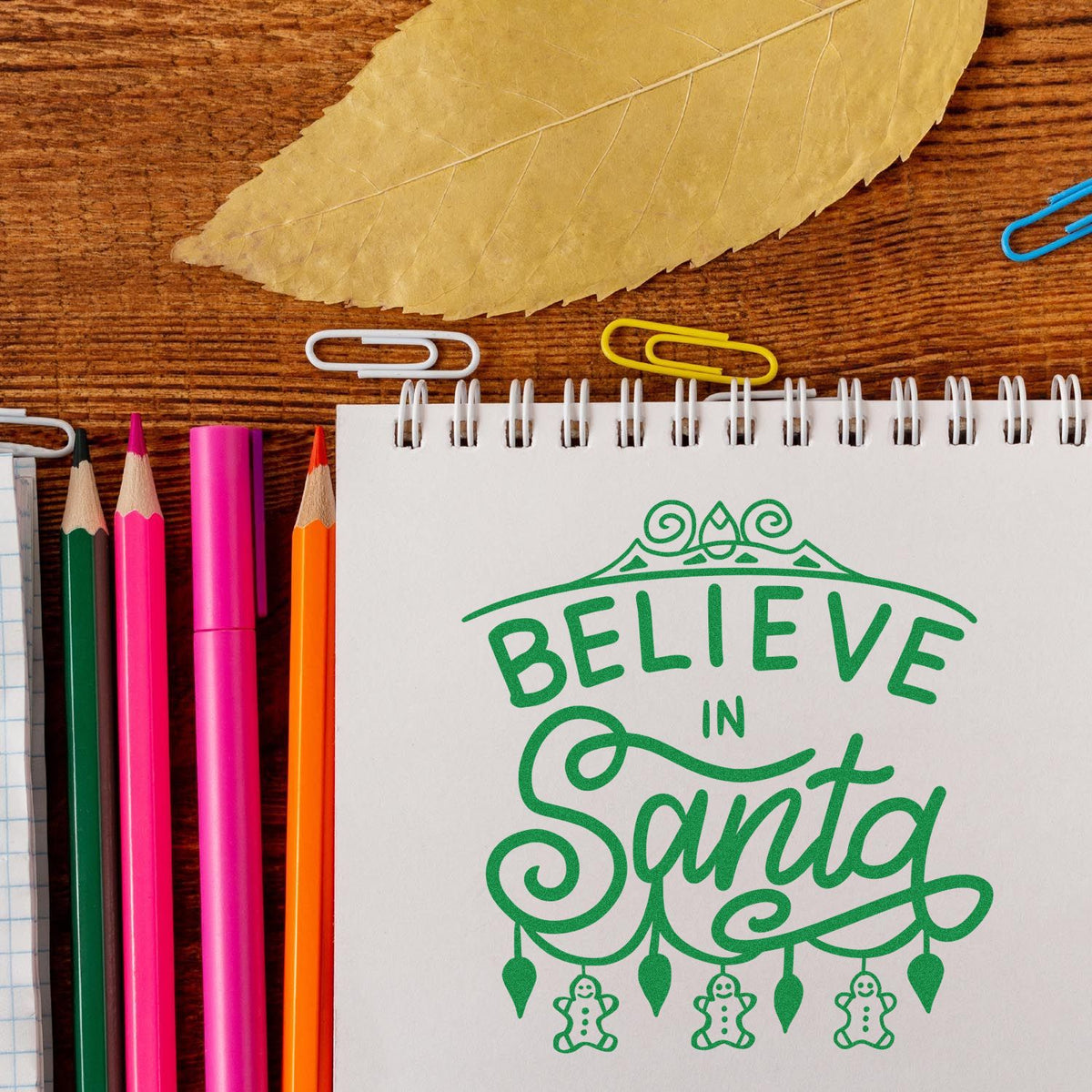 Christmas rubber stamp design reading Believe in Santa on a notebook, surrounded by colorful pencils and paper clips.