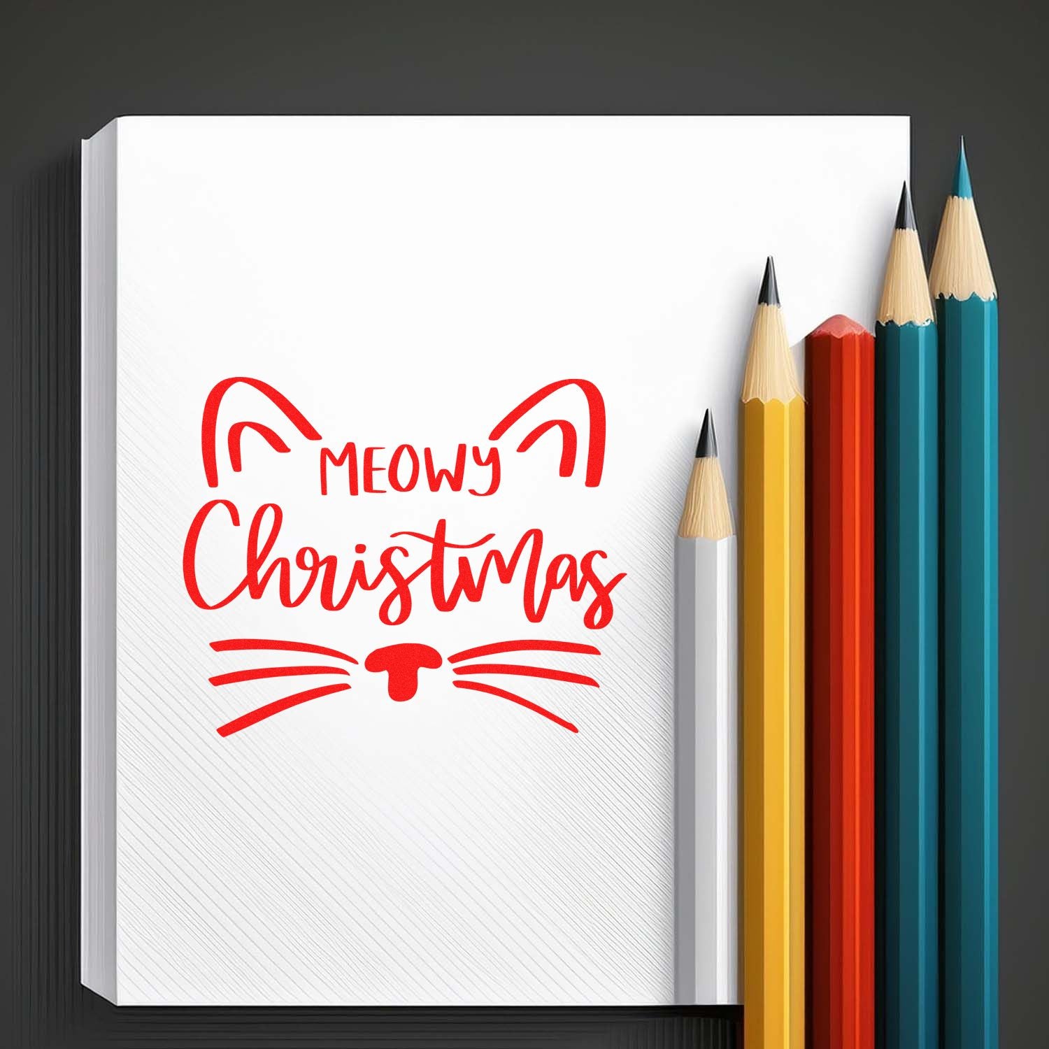 Meowy Christmas cat face rubber stamp design in red on notebook, with colored pencils for festive holiday crafting.