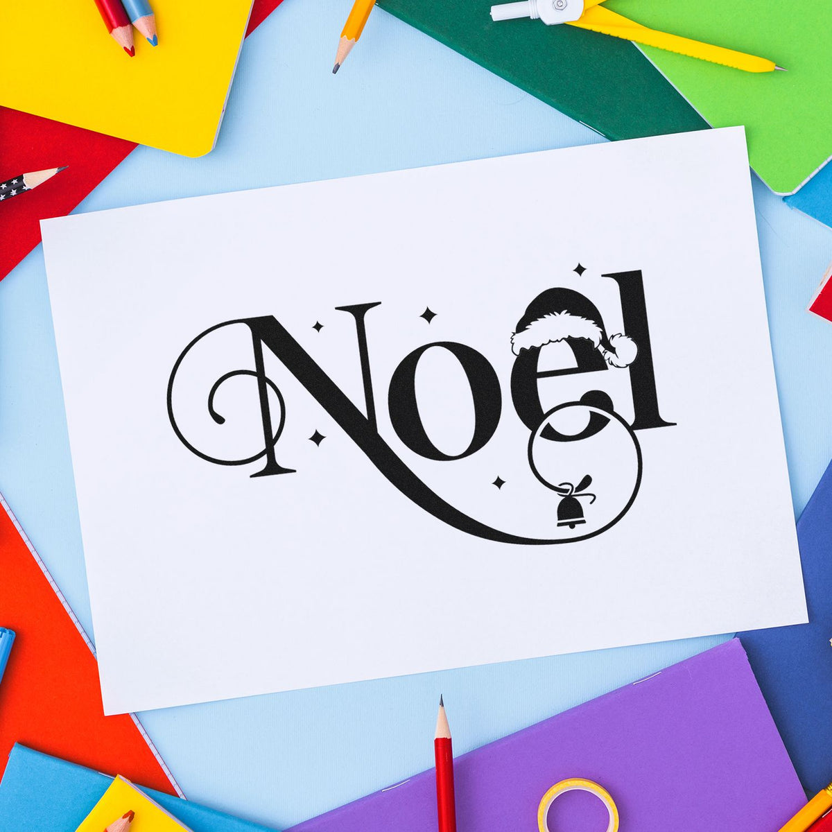 Christmas Noel rubber stamp design on white paper, surrounded by colorful stationery supplies for holiday card making.