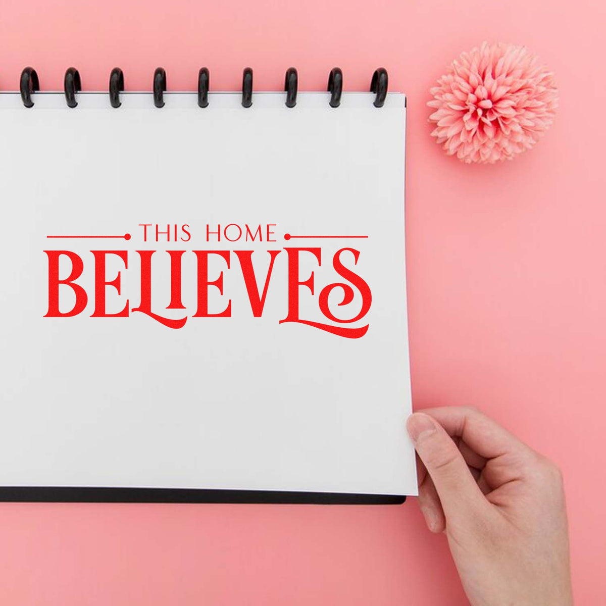 Custom rubber stamp mockup reading This Home Believes in red on a notebook with a pink background.
