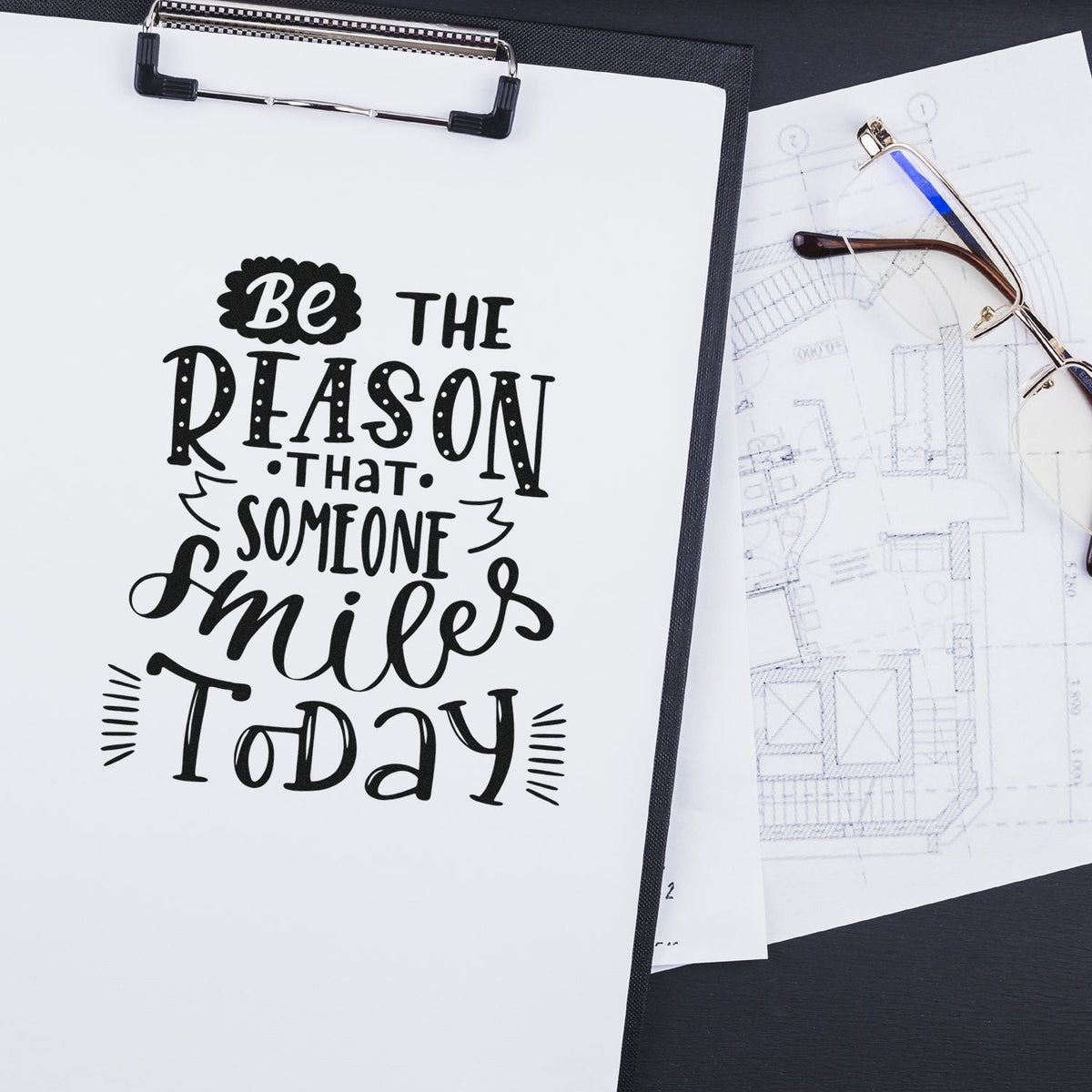 Motivational rubber stamp design with quote Be the reason that someone smiles today on clipboard beside eyeglasses and blueprint.