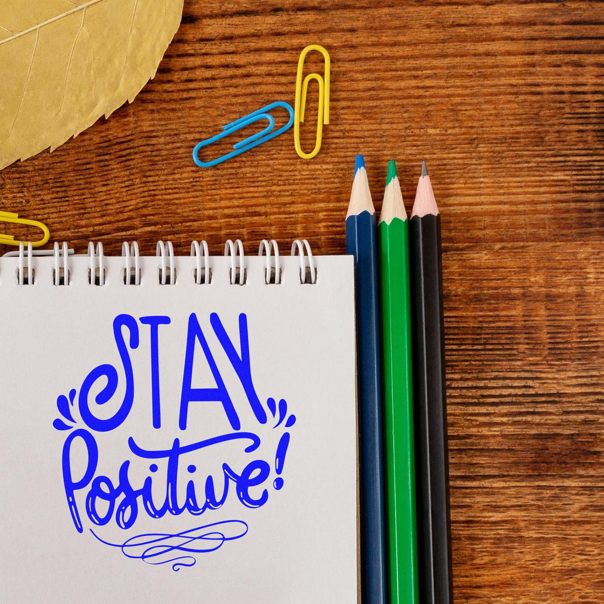 Motivational Stay Positive rubber stamp design on notebook, with pencils and paper clips on a wooden desk.
