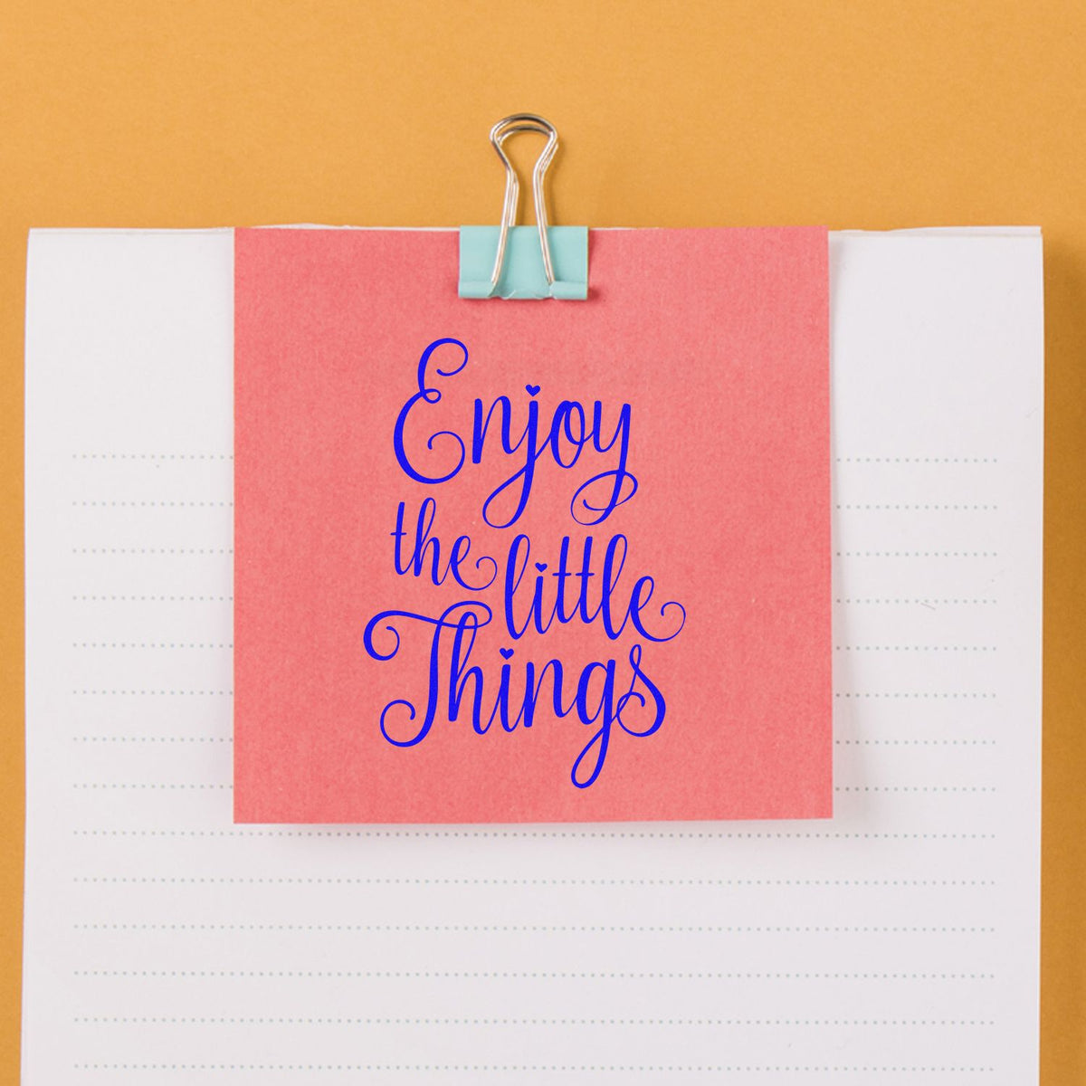Inspirational rubber stamp saying Enjoy the Little Things displayed on a pink note clipped to a notebook.