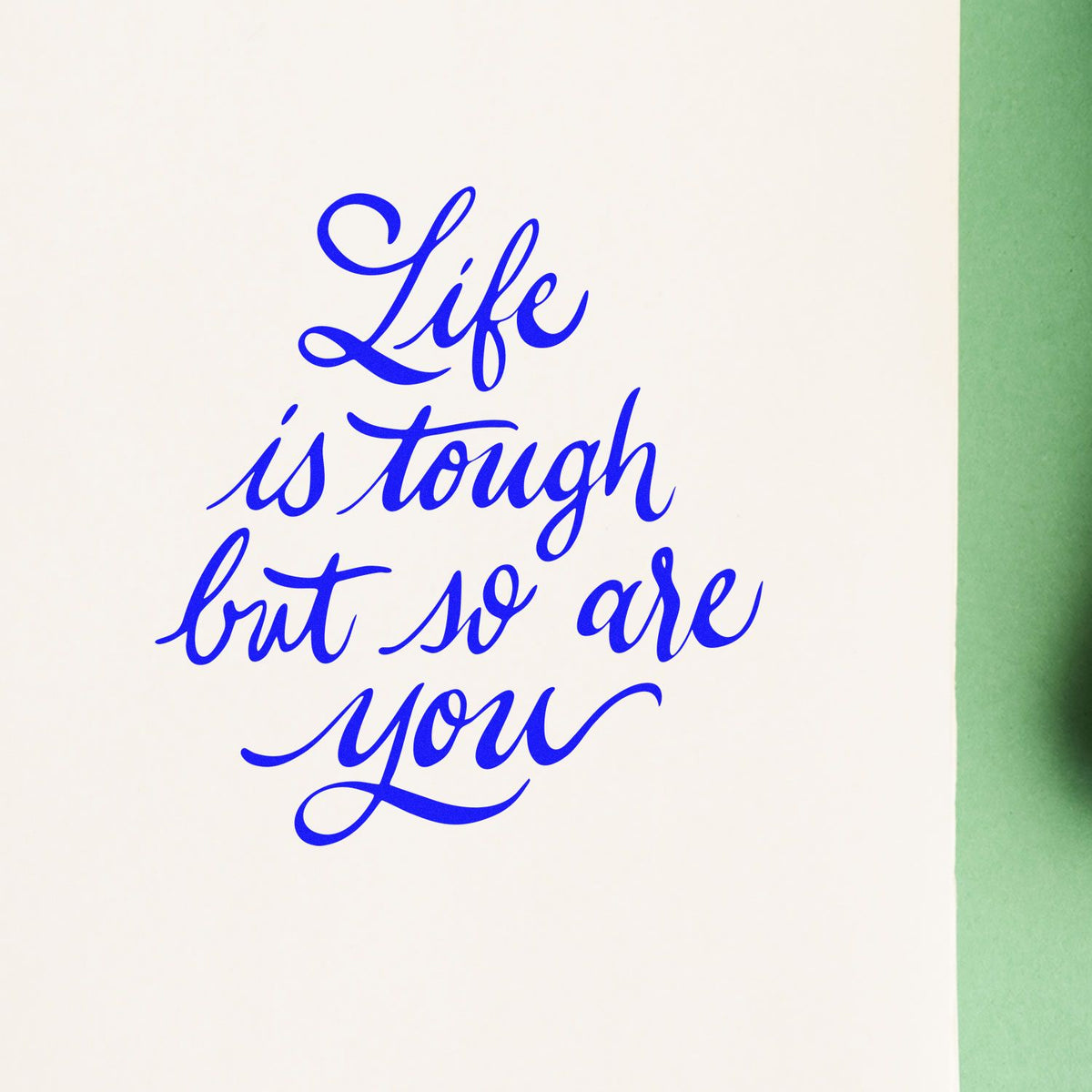 Custom rubber stamp in blue cursive with motivational quote, Life is tough but so are you, on white paper.