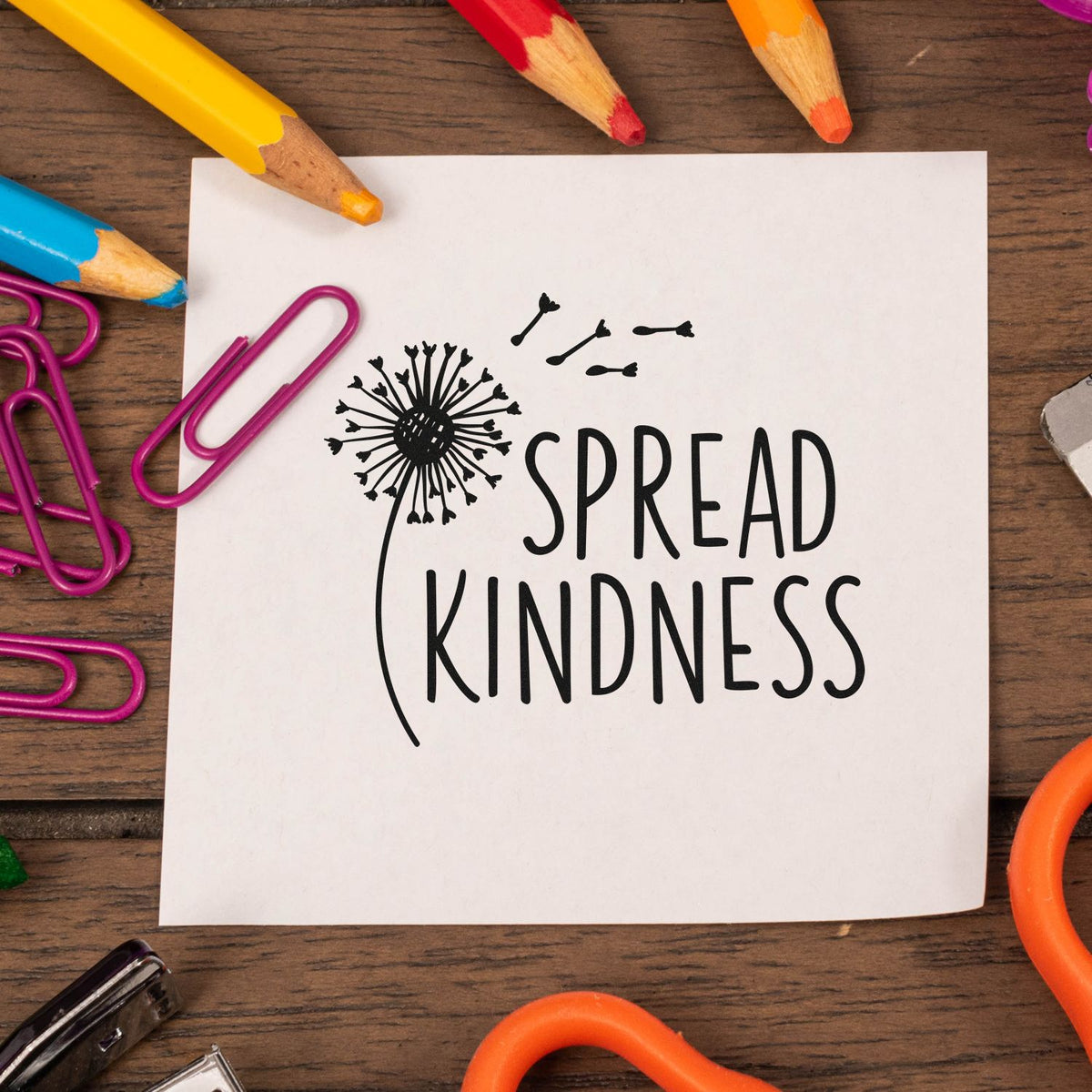 Spread Kindness rubber stamp design with dandelion and flying seeds, surrounded by pencils and paper clips on craft paper.