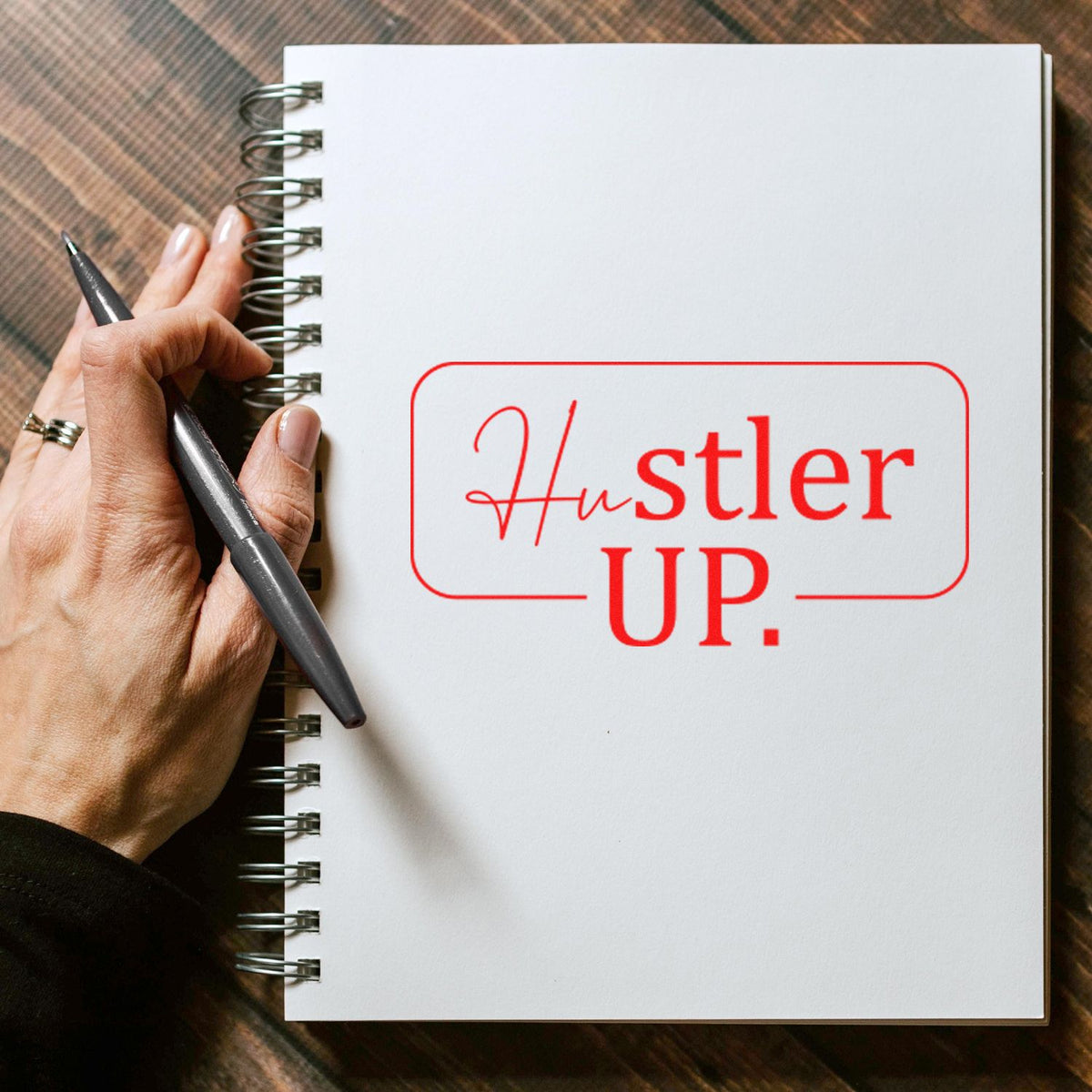 Custom rubber stamp reading Hustler Up on a spiral notebook, held by a hand with a pen for motivational branding.