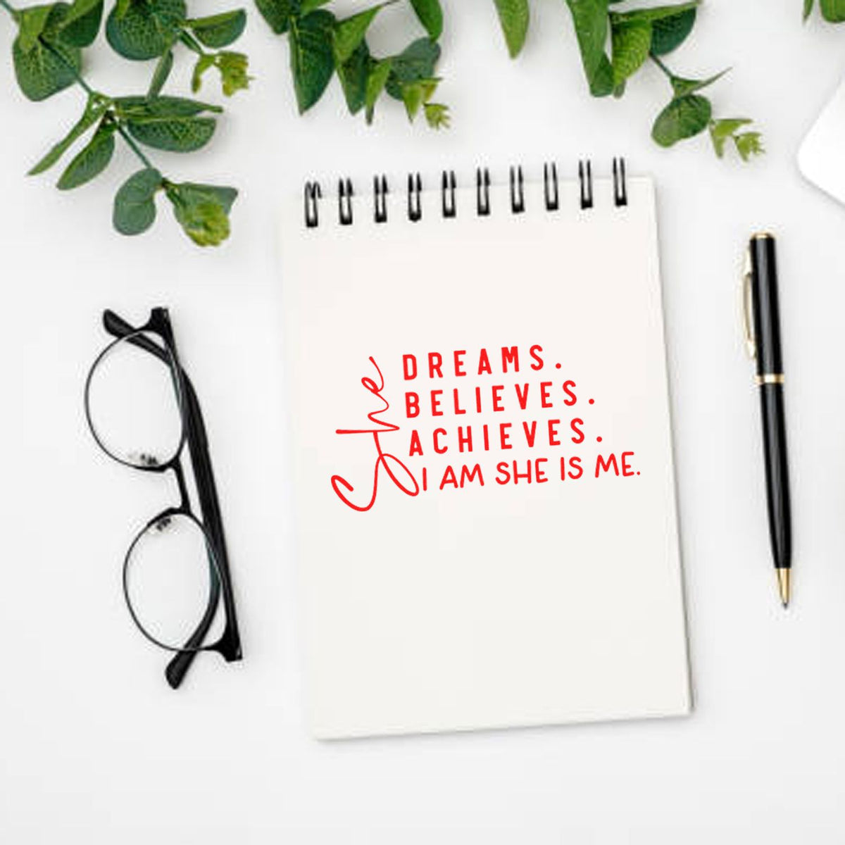 Motivational rubber stamp on notebook reading She dreams, believes, achieves. I am she is me with pen and glasses.