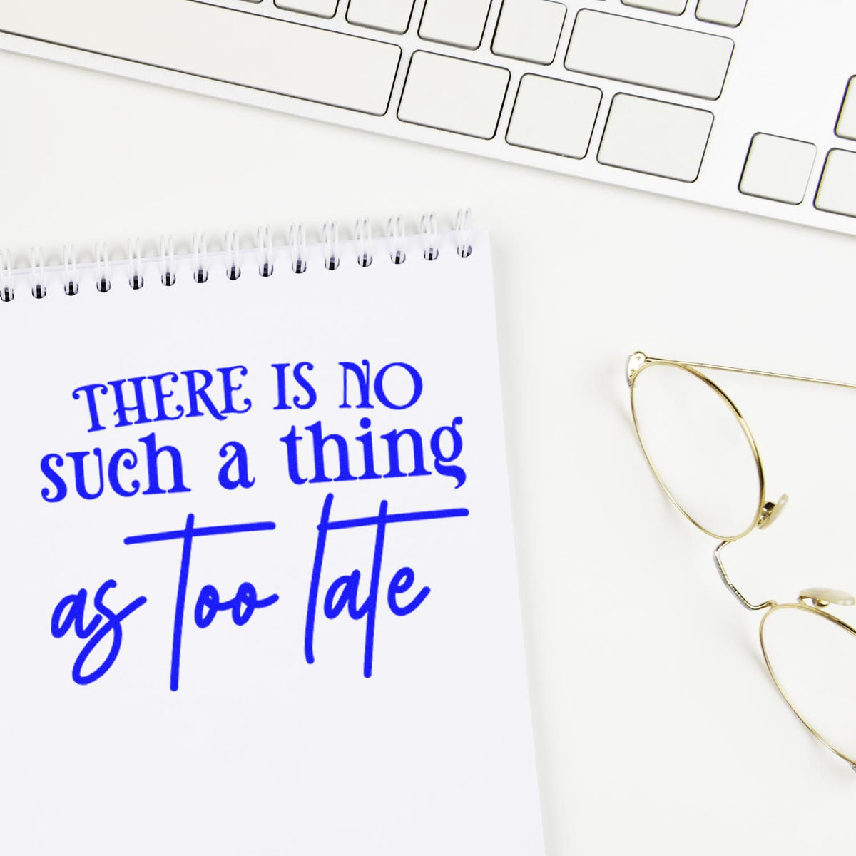 Motivational rubber stamp impression reading There is no such thing as too late on a notebook beside a keyboard and glasses.