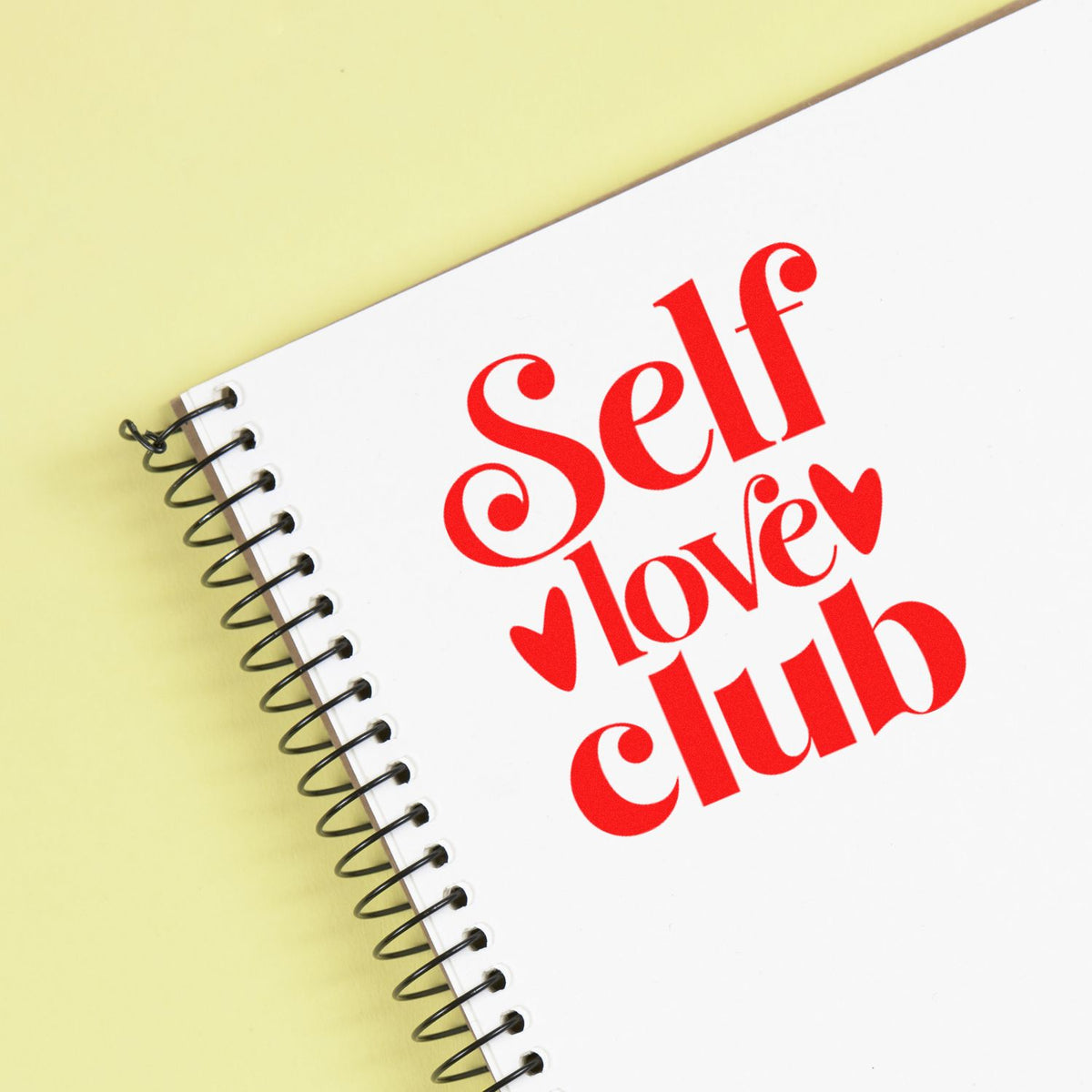 Self Love Club rubber stamp design on spiral notebook, featuring bold red lettering and hearts for self-care branding.