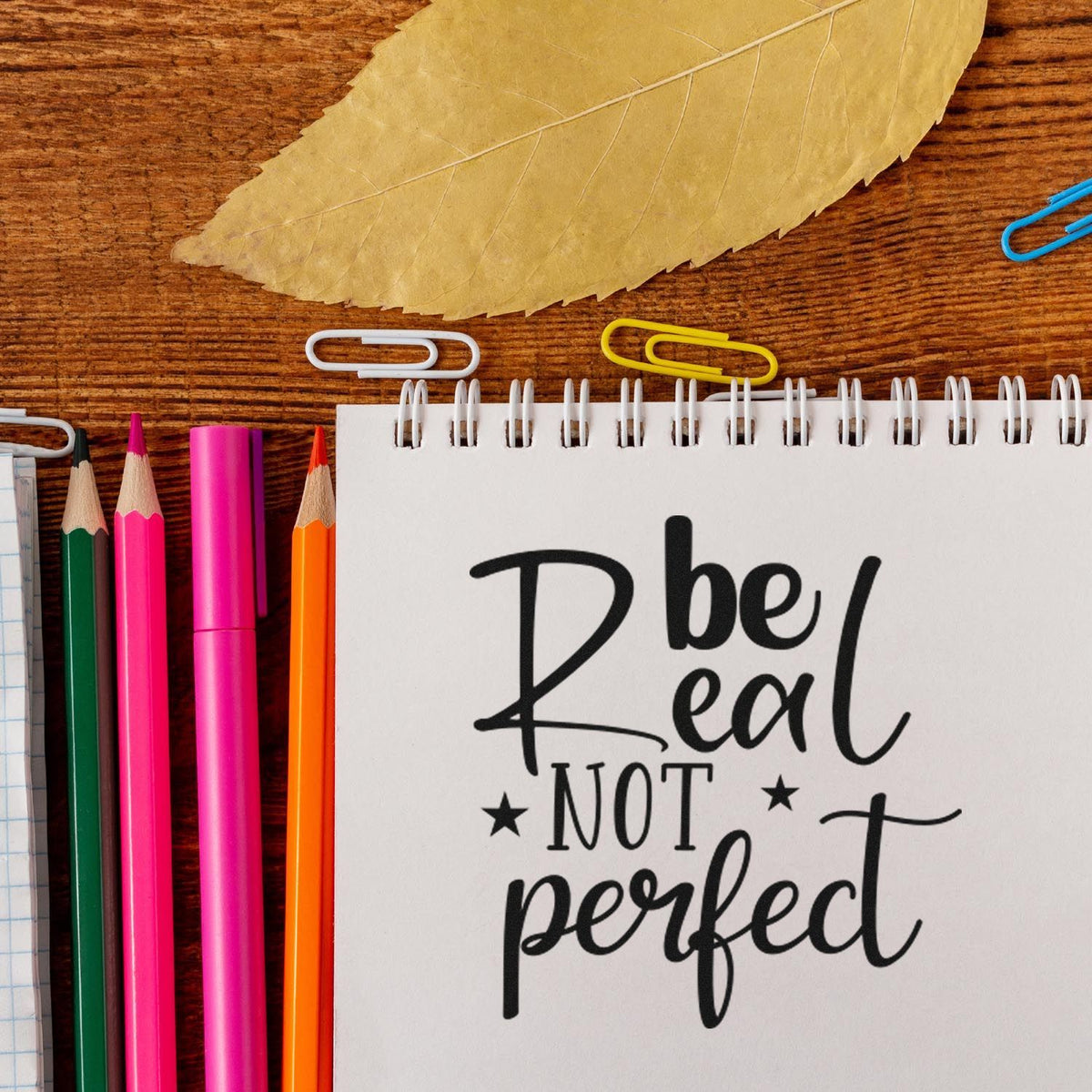 Motivational rubber stamp reading Be real not perfect in a notebook, with colorful pencils and paper clips on a wooden desk.