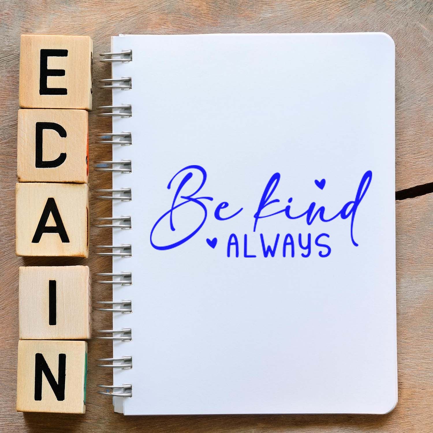 Inspirational rubber stamp reading Be Kind Always in blue on a spiral notebook, with wooden letter blocks beside it.
