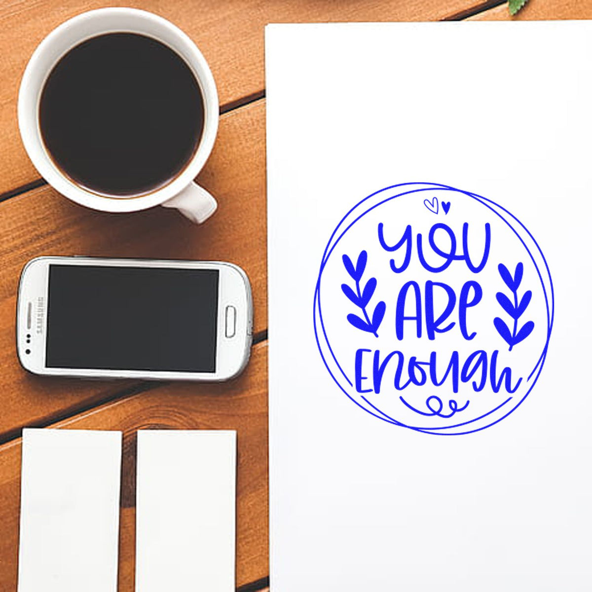 Custom rubber stamp with blue ink reading You Are Enough on paper, shown on a wooden desk with coffee and phone.