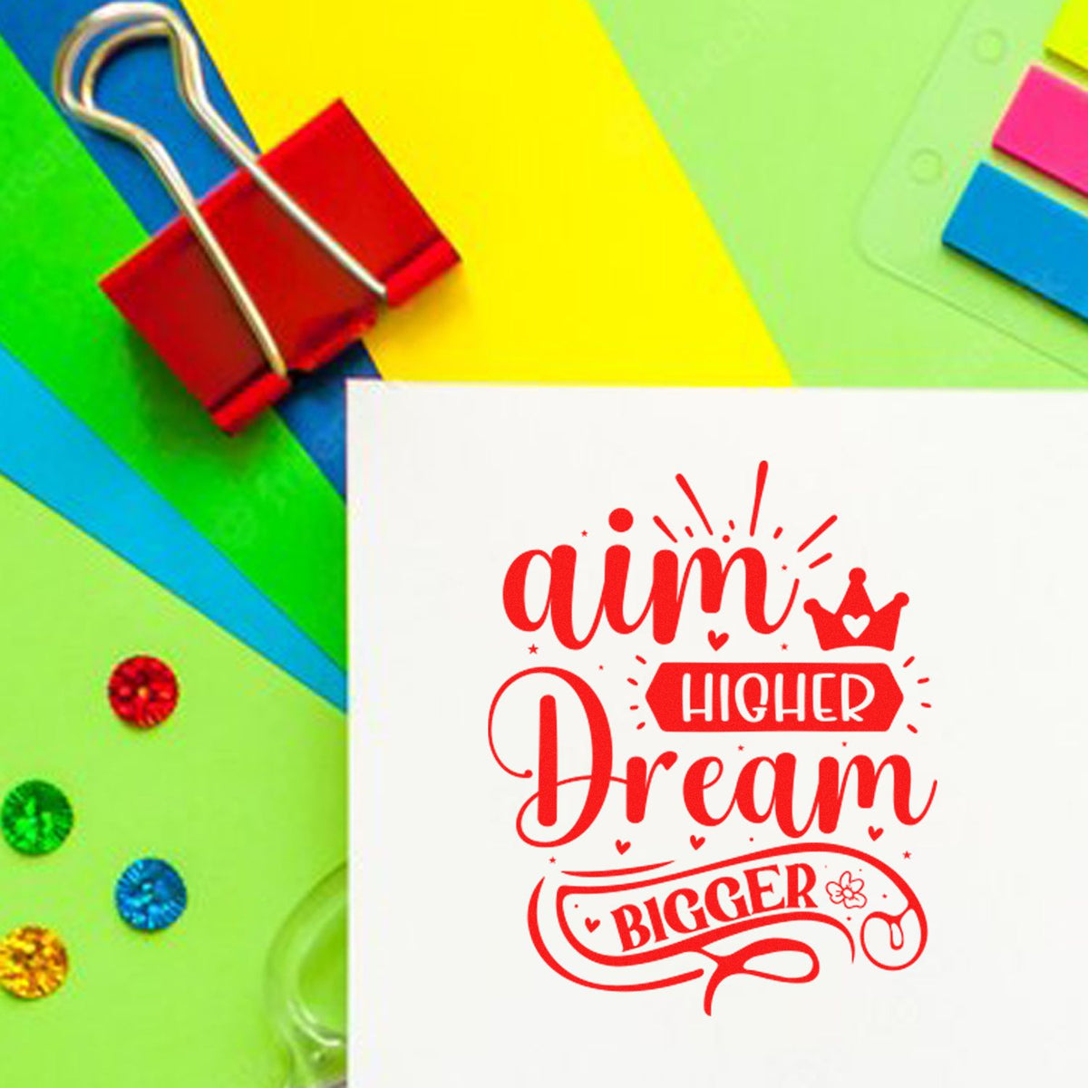 Motivational rubber stamp design with Aim Higher Dream Bigger in red on paper with colorful stationery background.