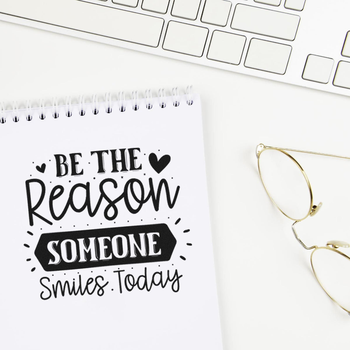 Motivational rubber stamp with Be the reason someone smiles today quote on notebook beside keyboard and glasses.
