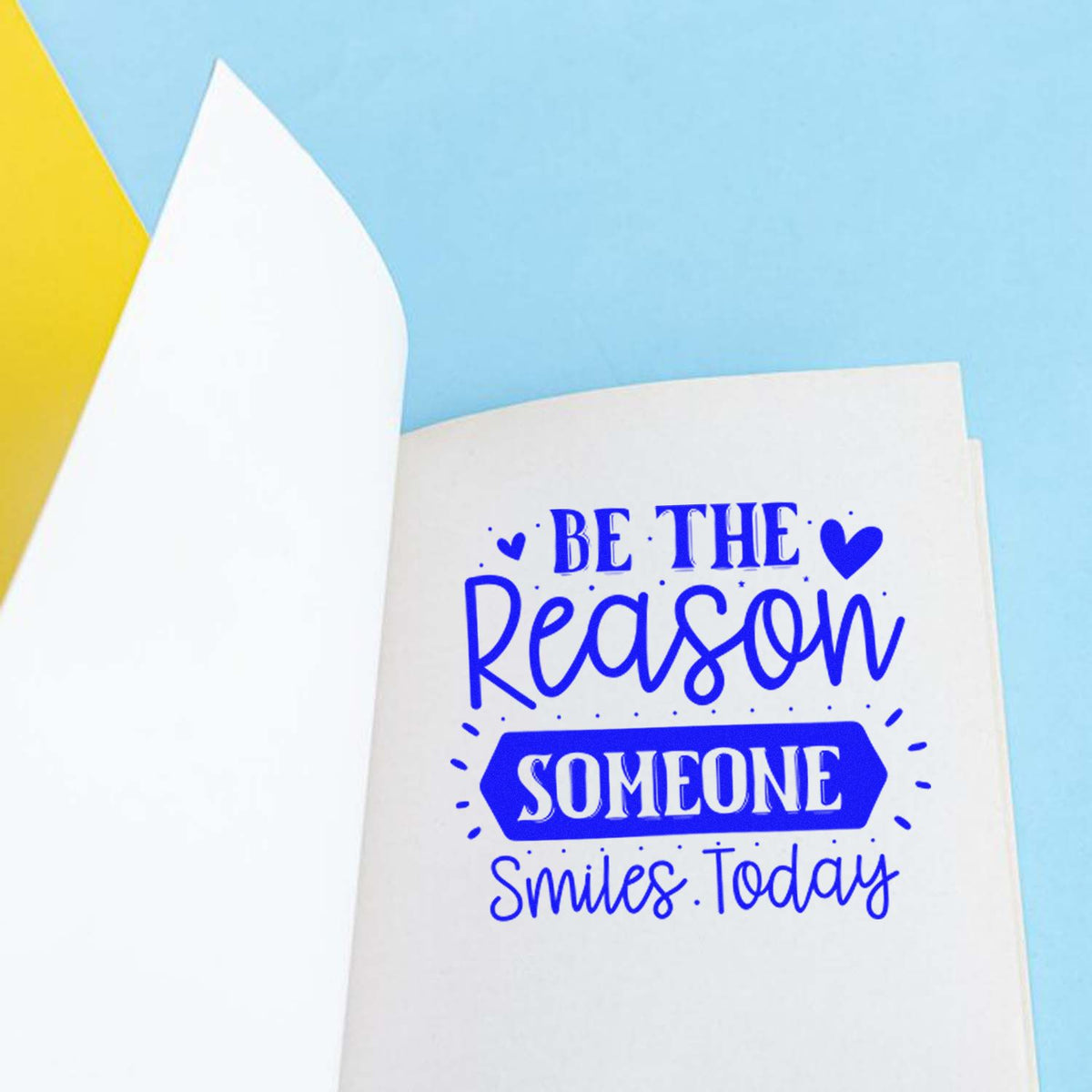 Motivational rubber stamp saying Be the reason someone smiles today in blue on paper.