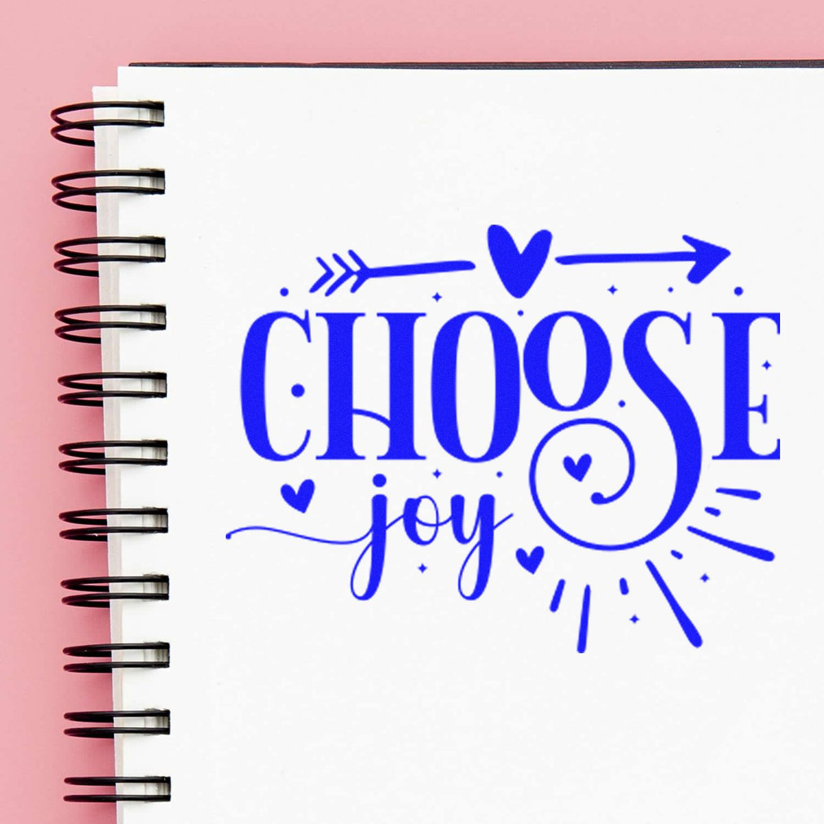 Blue Choose Joy rubber stamp impression with hearts and arrows on a spiral notebook.