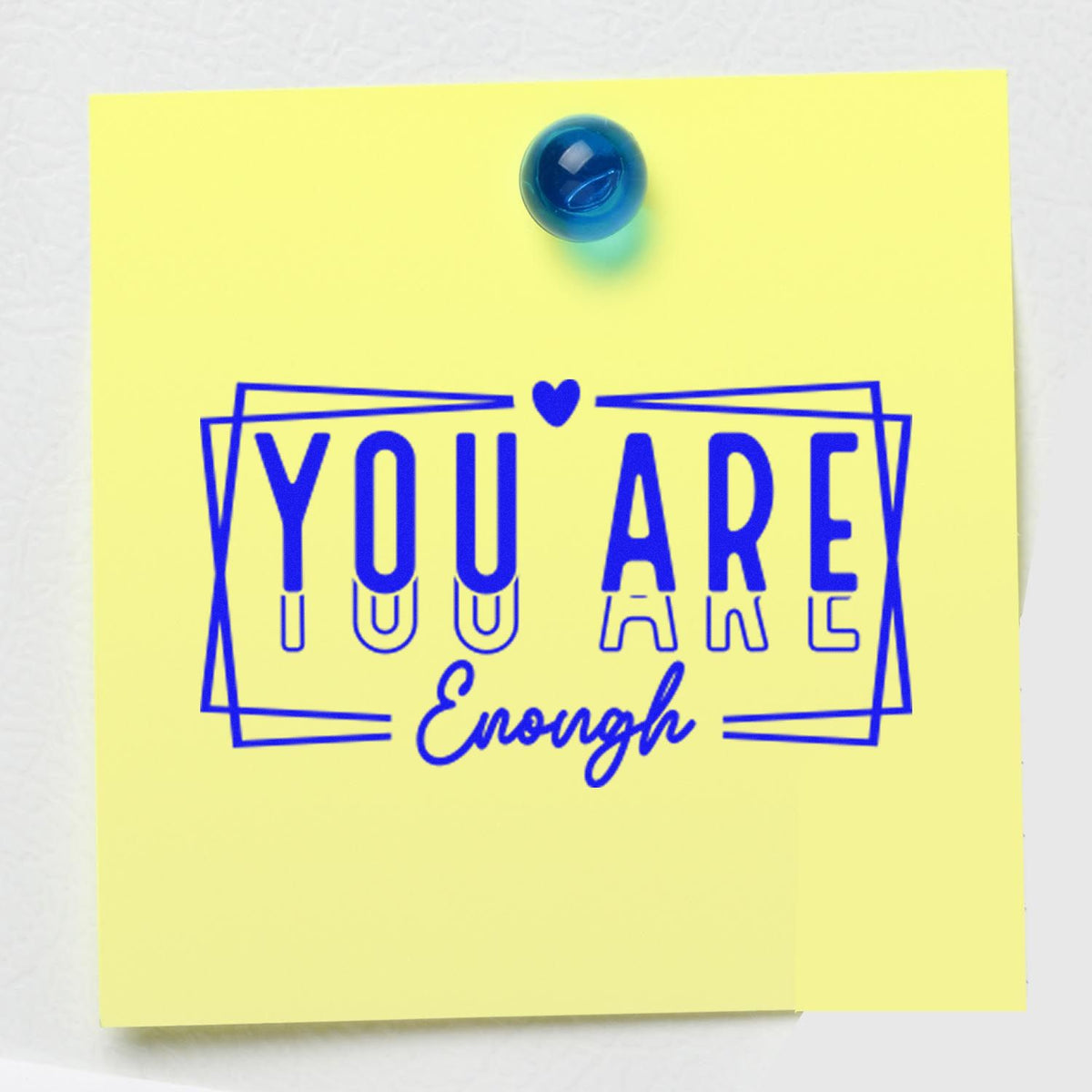 Motivational rubber stamp reading You Are Enough in blue on a yellow sticky note, pinned with a blue tack.
