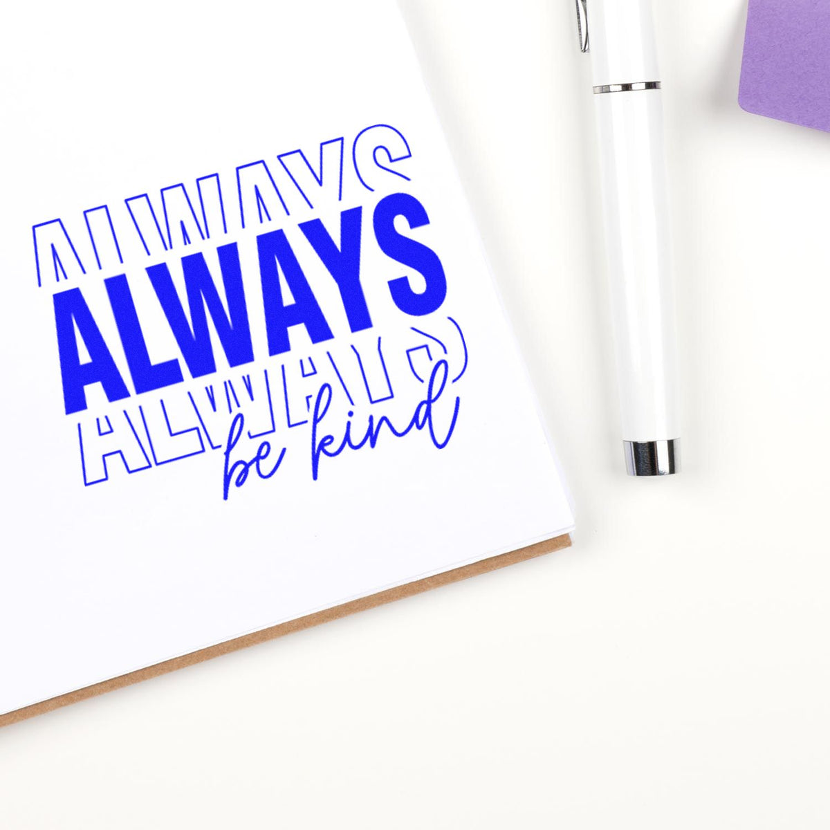 Blue rubber stamp design reading Always be kind on white paper with pen, showcasing a custom motivational rubber stamp.