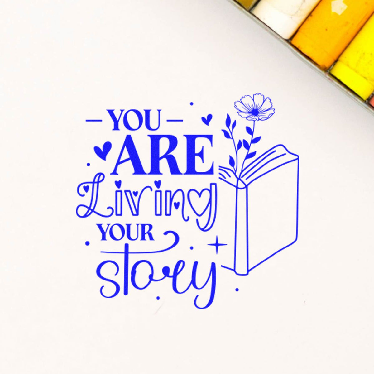Blue rubber stamp reading You Are Living Your Story with book and flower design on paper.