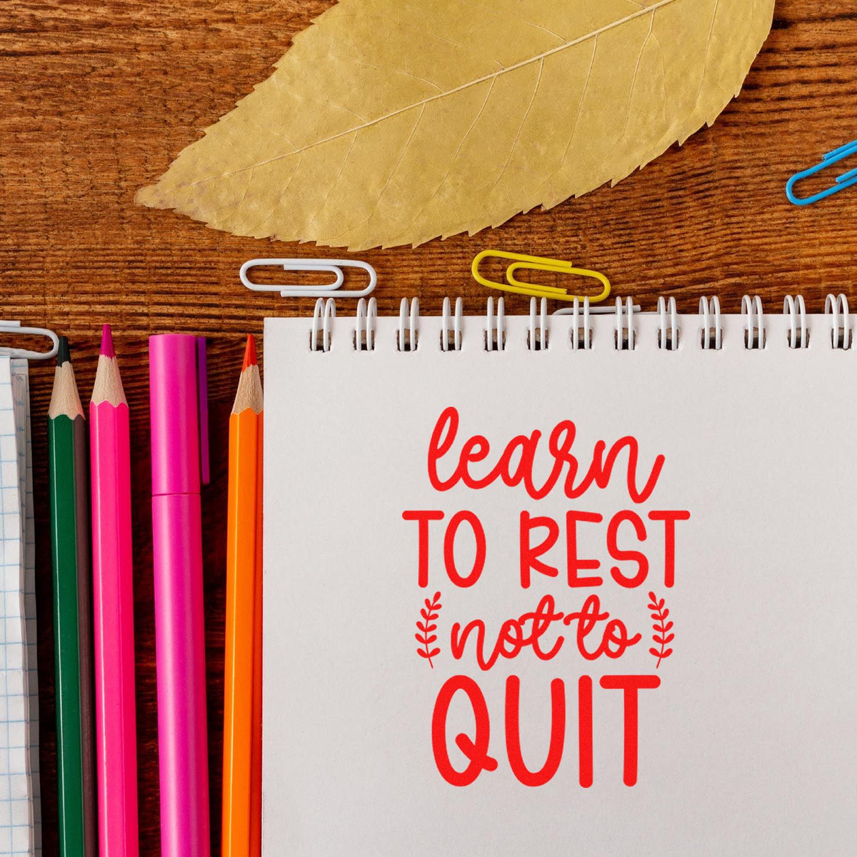 Motivational rubber stamp with Learn to rest, not to quit on a notebook, styled with pencils, paper clips and leaf.