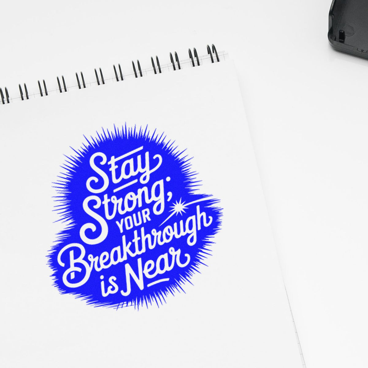 Motivational rubber stamp impression reading Stay Strong, Your Breakthrough Is Near on a notebook page.