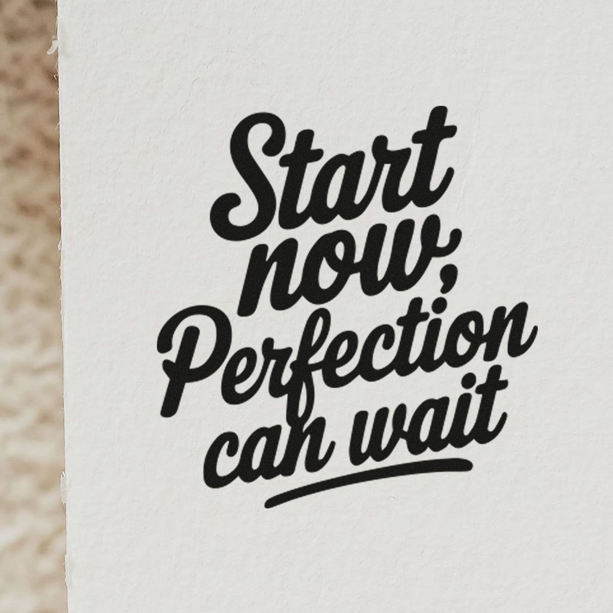 Motivational rubber stamp imprint reading Start now, perfection can wait in bold script on white paper.