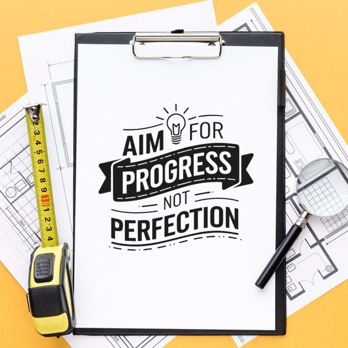 Motivational rubber stamp design reading Aim for progress, not perfection on clipboard with blueprint and measuring tools.