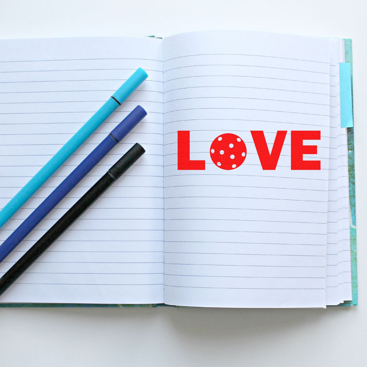 Red LOVE rubber stamp on lined notebook with colorful pens, perfect for handmade cards, journaling, and craft projects.