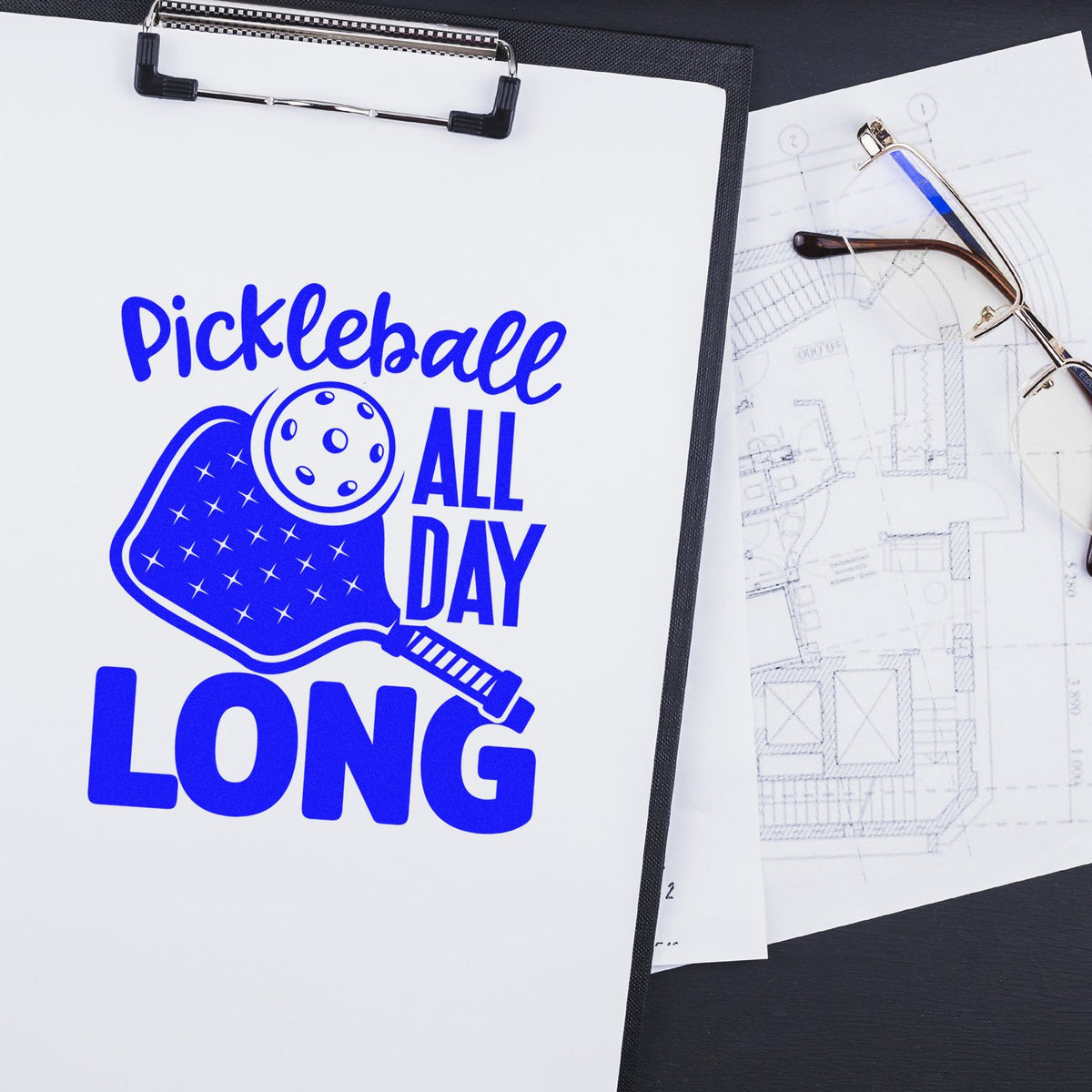 Pickleball All Day Long custom rubber stamp design with paddle and ball icon on a clipboard next to blueprint and glasses.