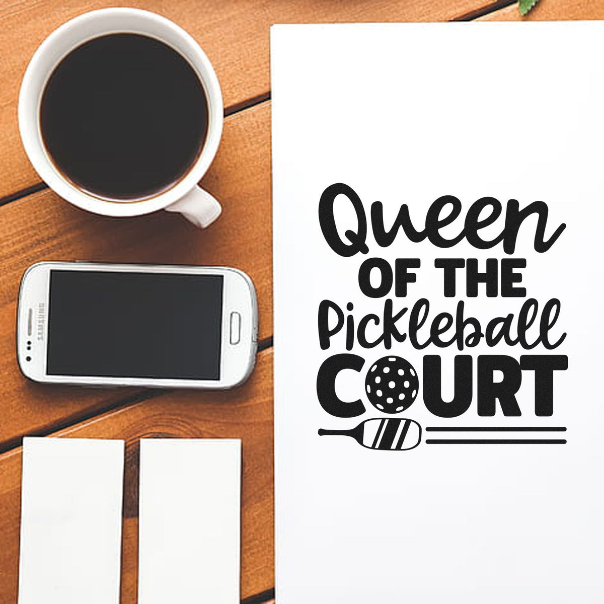Queen of the Pickleball Court rubber stamp design mockup with paddle and ball graphic on paper beside coffee and phone