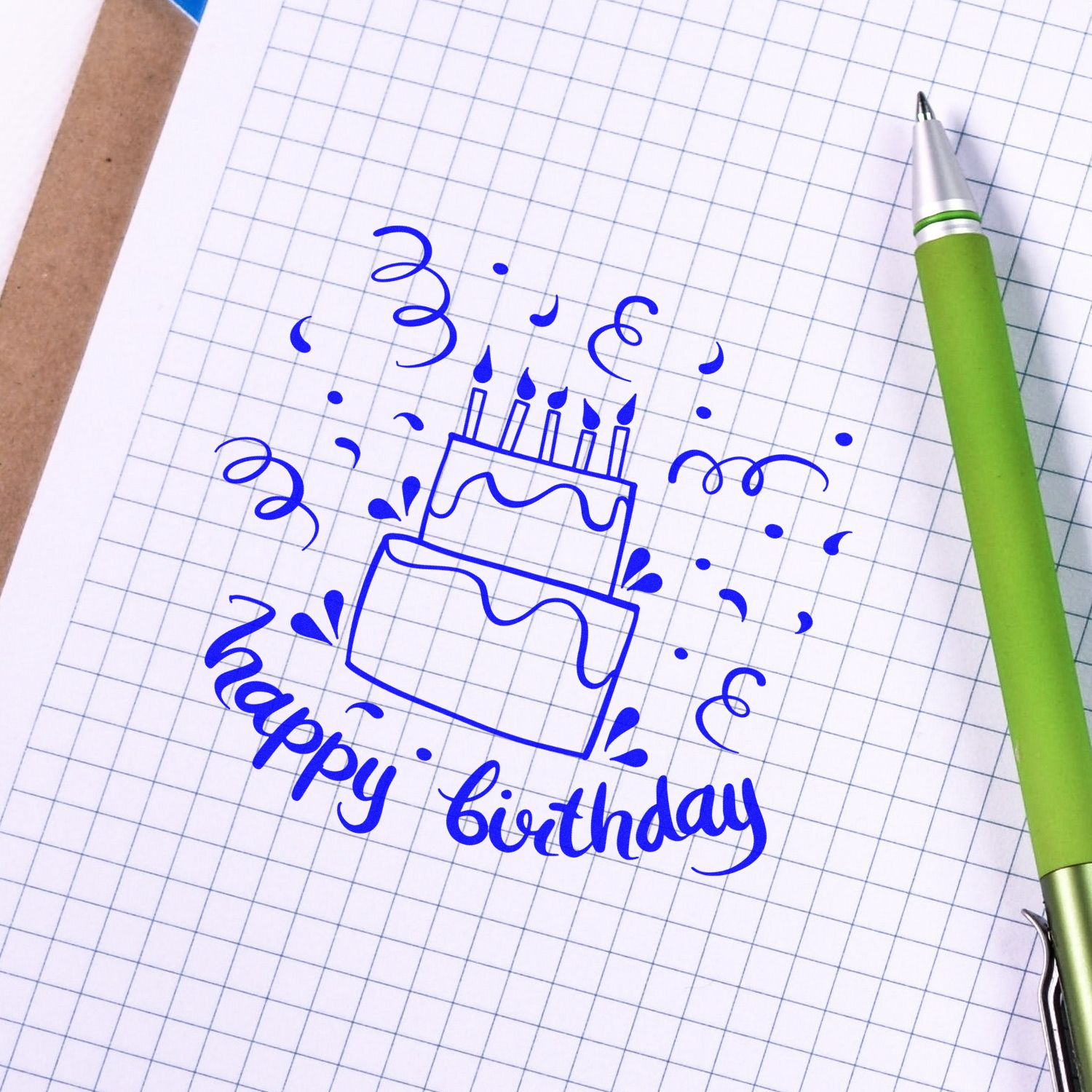 Happy birthday rubber stamp design with cake icon on graph paper, beside a green pen.