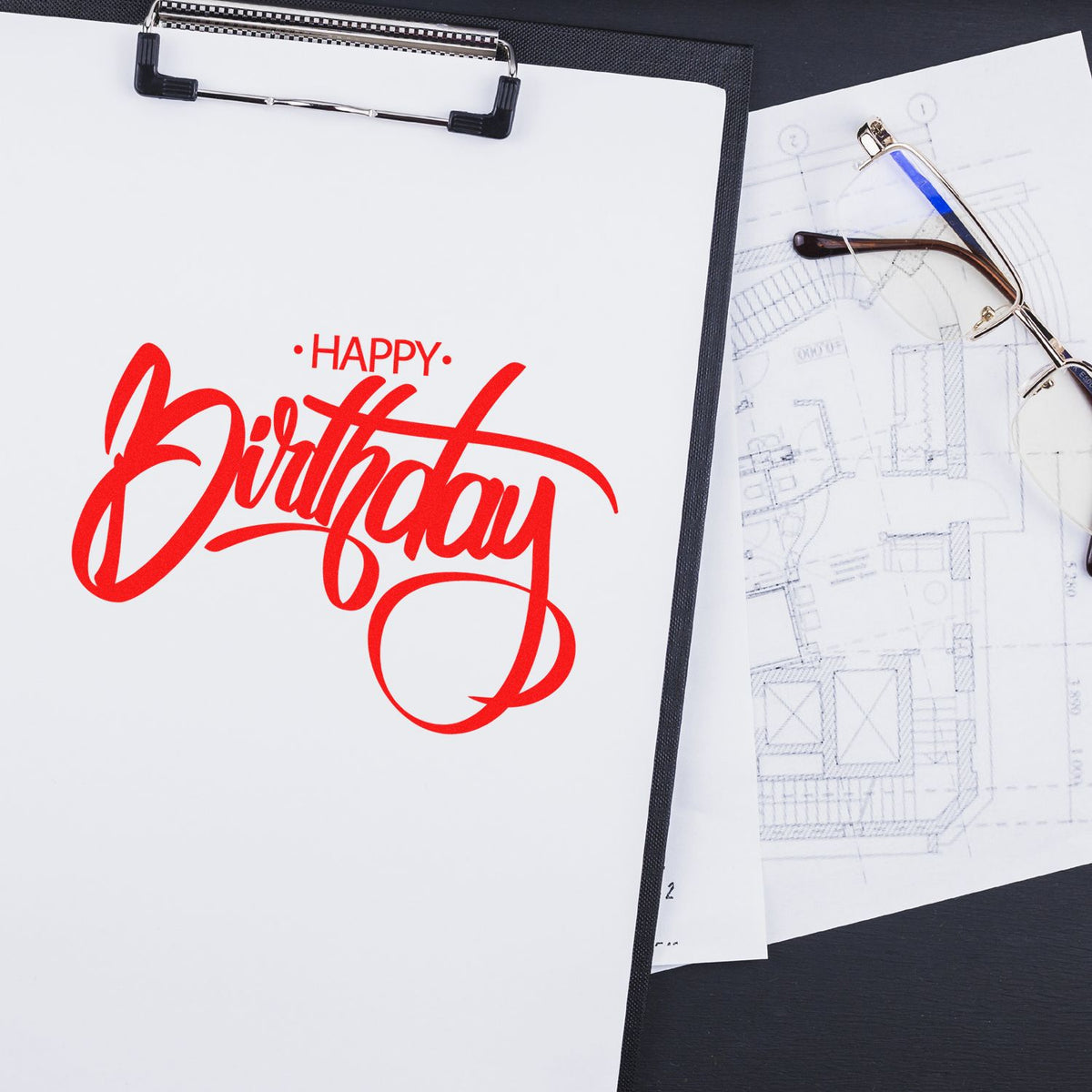 Custom Happy Birthday rubber stamp design in red script on clipboard, with blueprint sketches and eyeglasses.