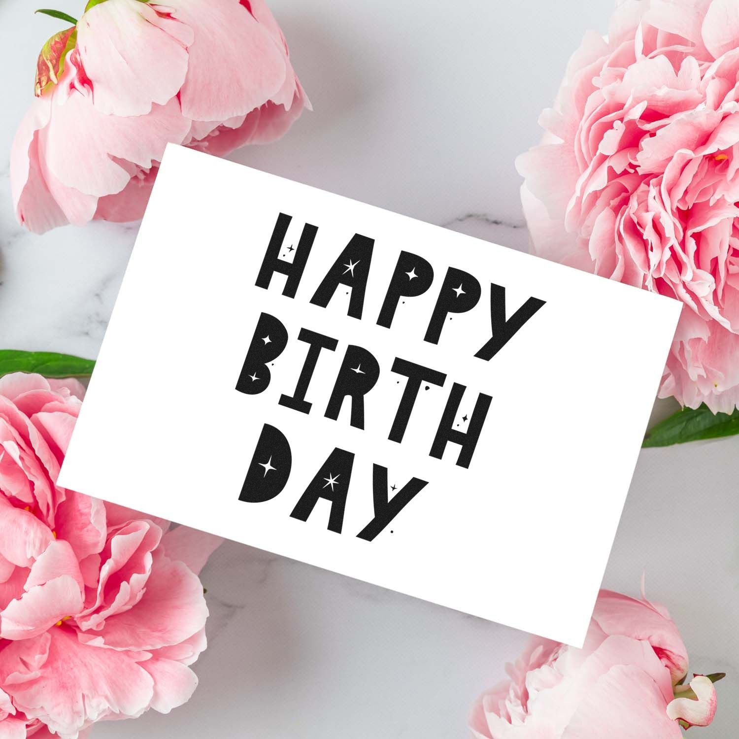 Happy Birthday rubber stamp with bold black text and star accents, framed by pink peonies on marble.