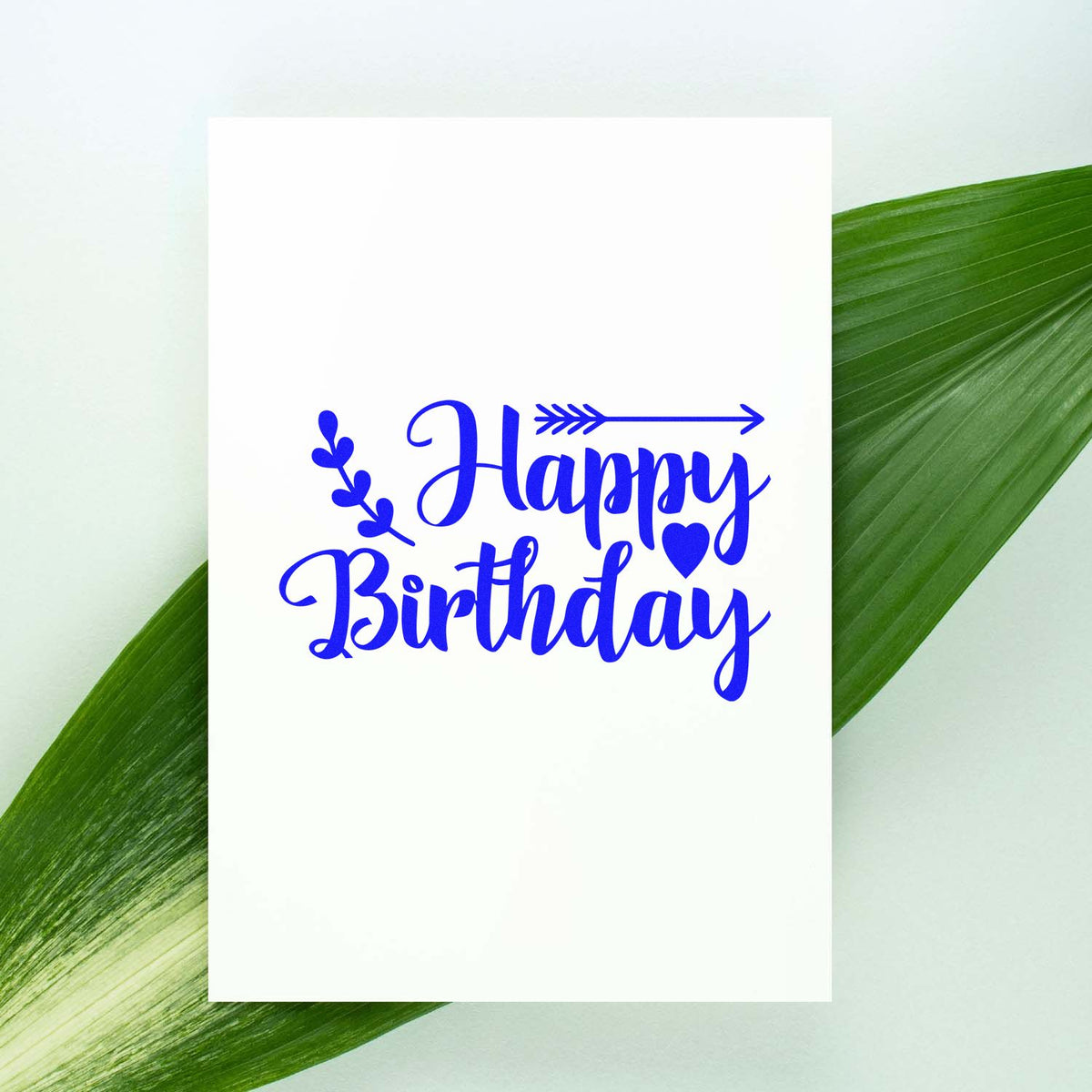 Happy Birthday rubber stamp design with blue script, heart, arrow and leaf accents on a greeting card mockup.