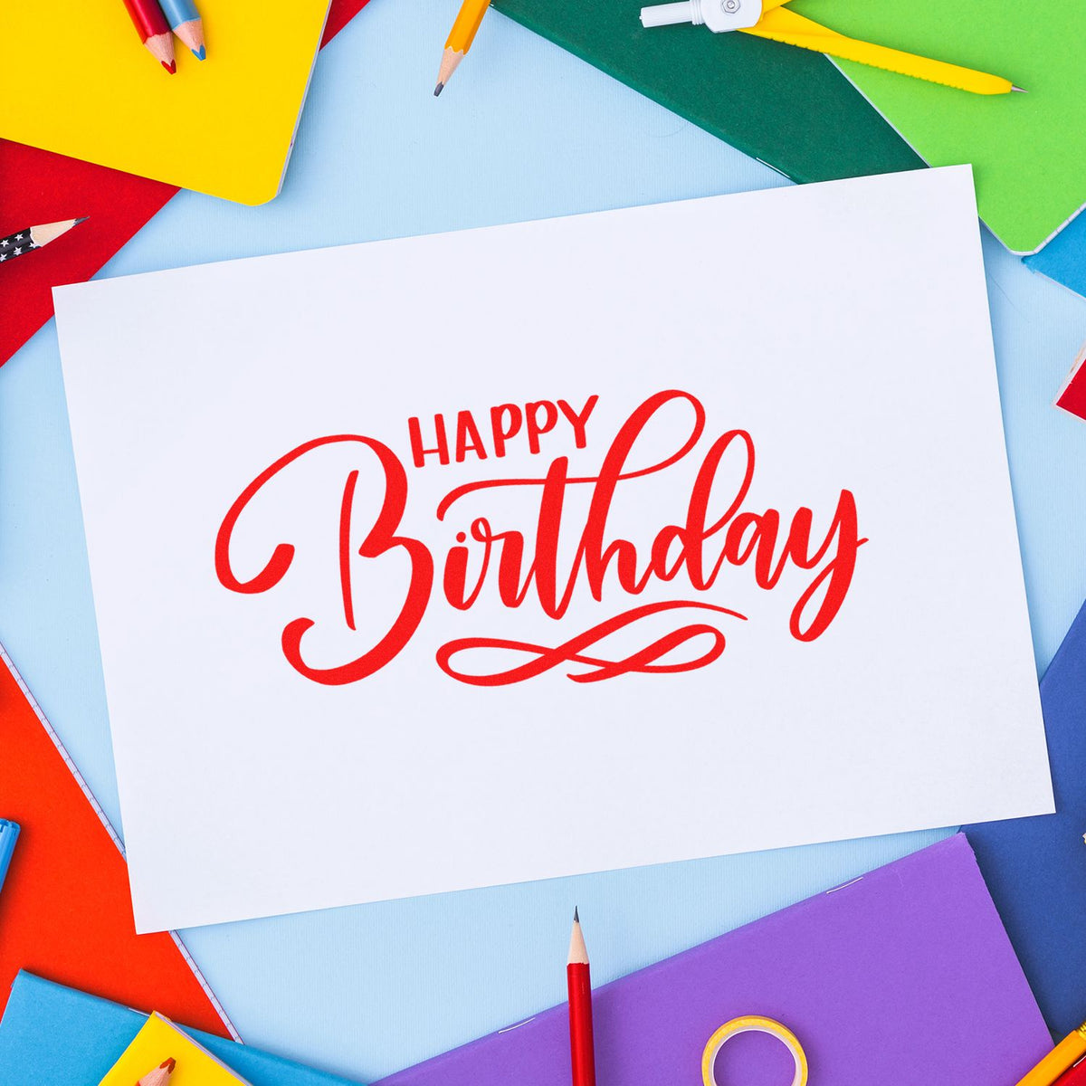 Happy Birthday rubber stamp design in red script on white paper, surrounded by colorful pencils and notebooks.