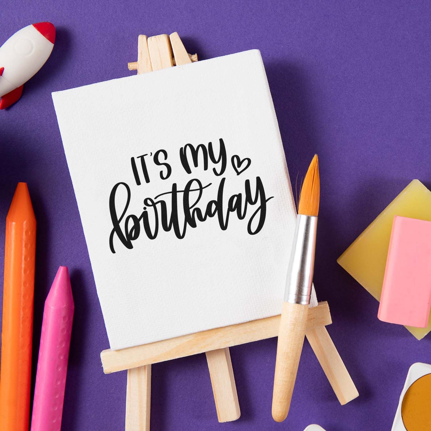 Birthday rubber stamp design reading Its my birthday displayed on a mini easel with art supplies on a purple background.