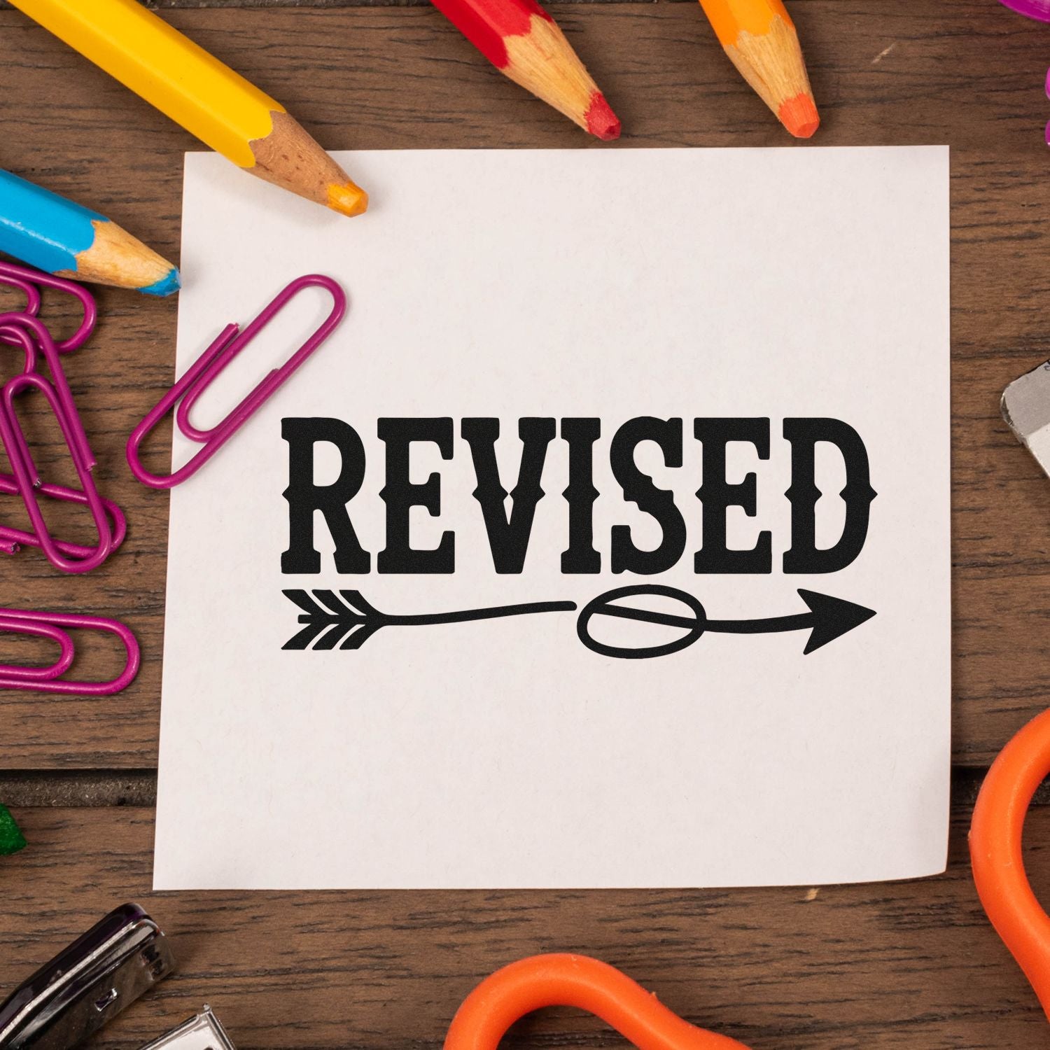 Revised rubber stamp design on paper with arrow graphic, surrounded by colored pencils and paper clips on a wooden desk.