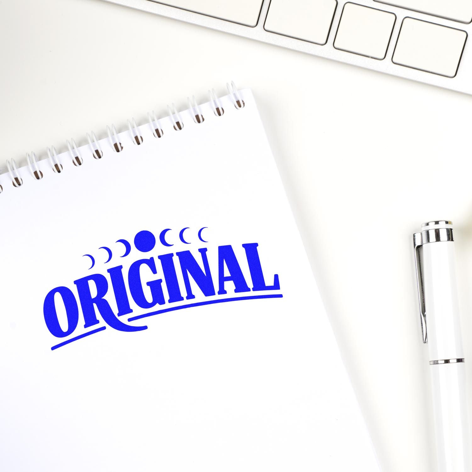 Blue ORIGINAL rubber stamp on a notebook beside a pen and keyboard, suggesting document approval or authenticity.