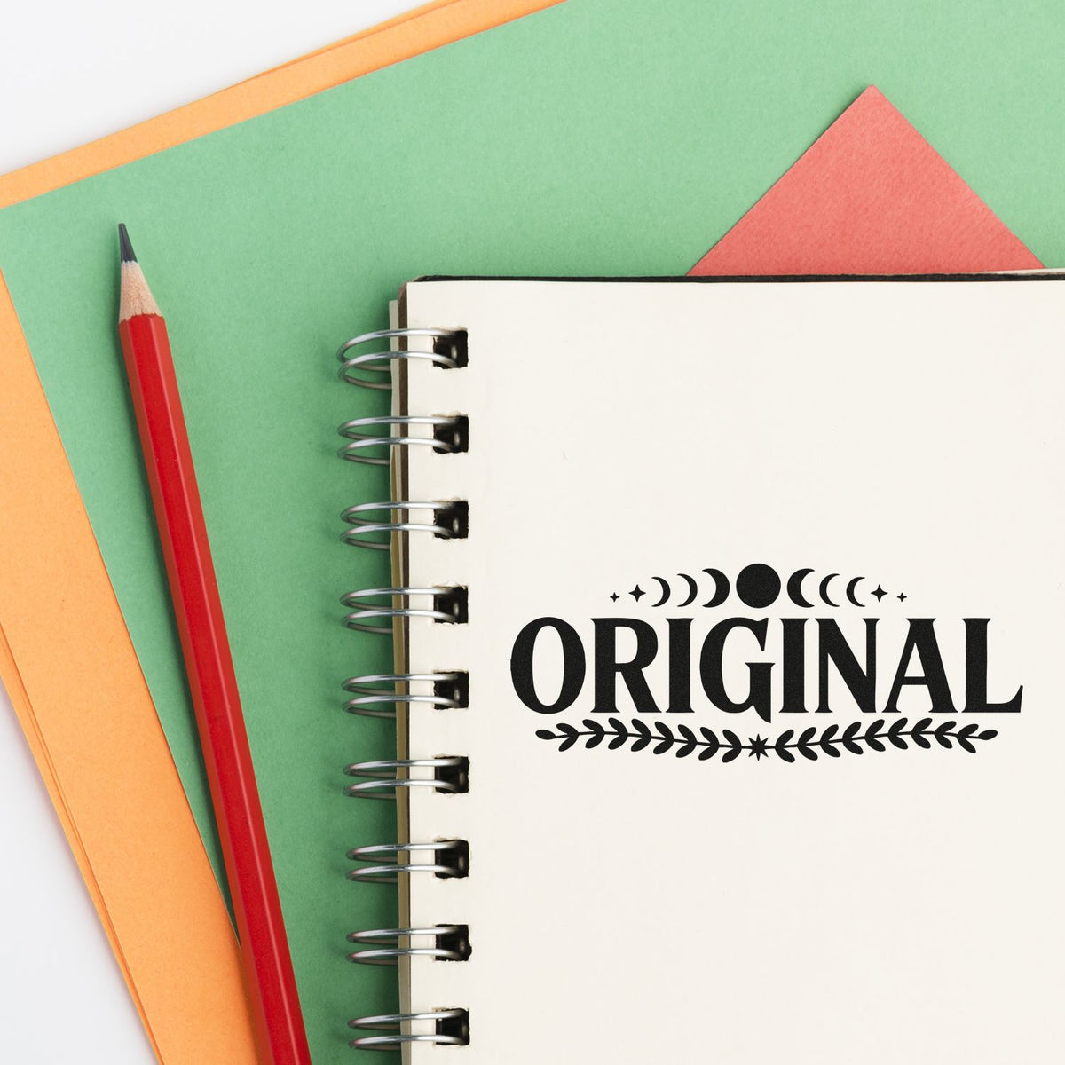 Custom rubber stamp reading Original on a spiral notebook, with pencil and colorful paper background.
