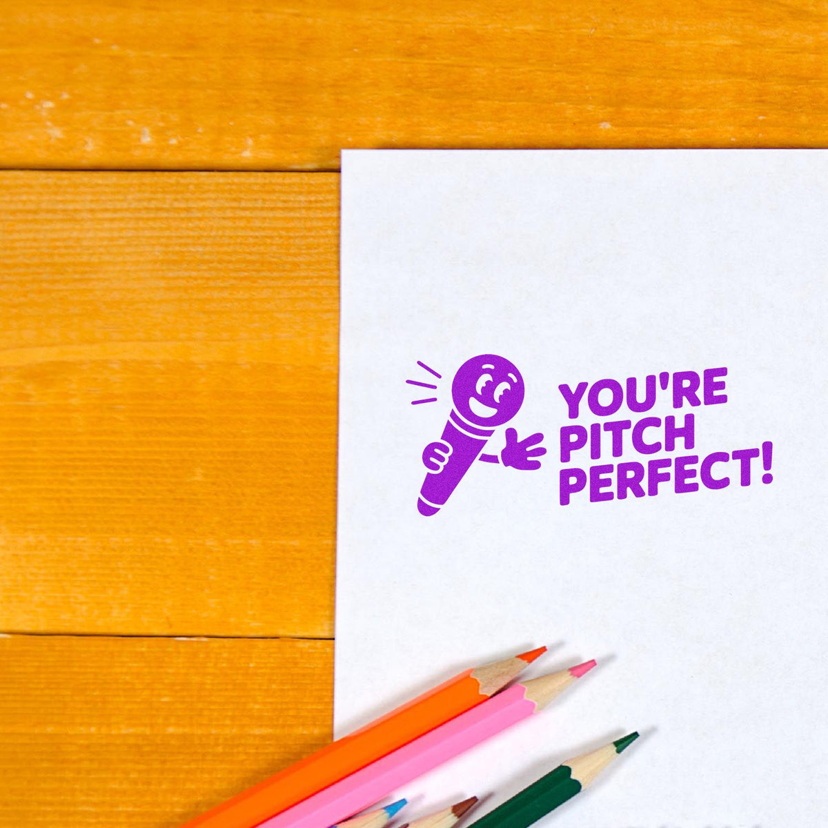 Purple rubber stamp saying Youre Pitch Perfect! with microphone icon on paper, shown with colored pencils on a wooden desk.