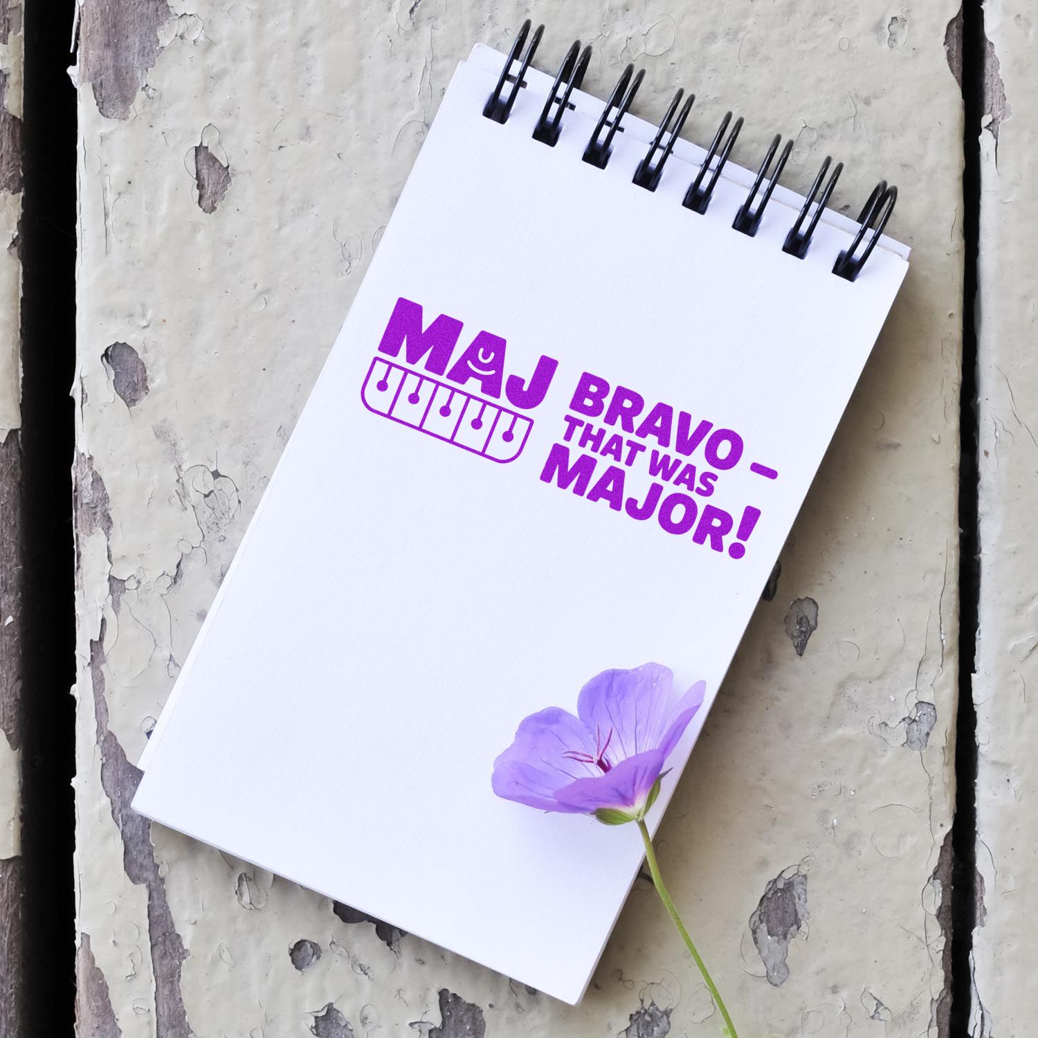 Custom rubber stamp imprint reading Maj Bravo That Was Major! on a notebook with a purple flower accent.