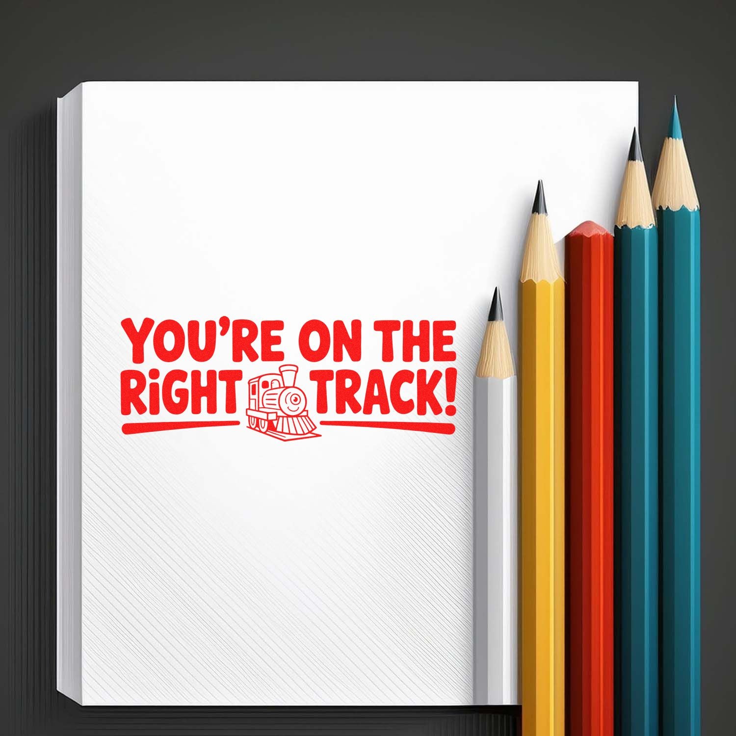 Rubber stamp design reading Youre on the right track! with train icon on a notebook page, surrounded by colored pencils.