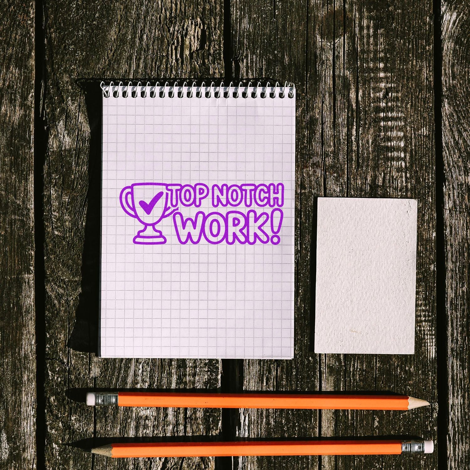Top Notch Work! rubber stamp impression with trophy icon on graph notebook, beside blank card and pencils on wood table.