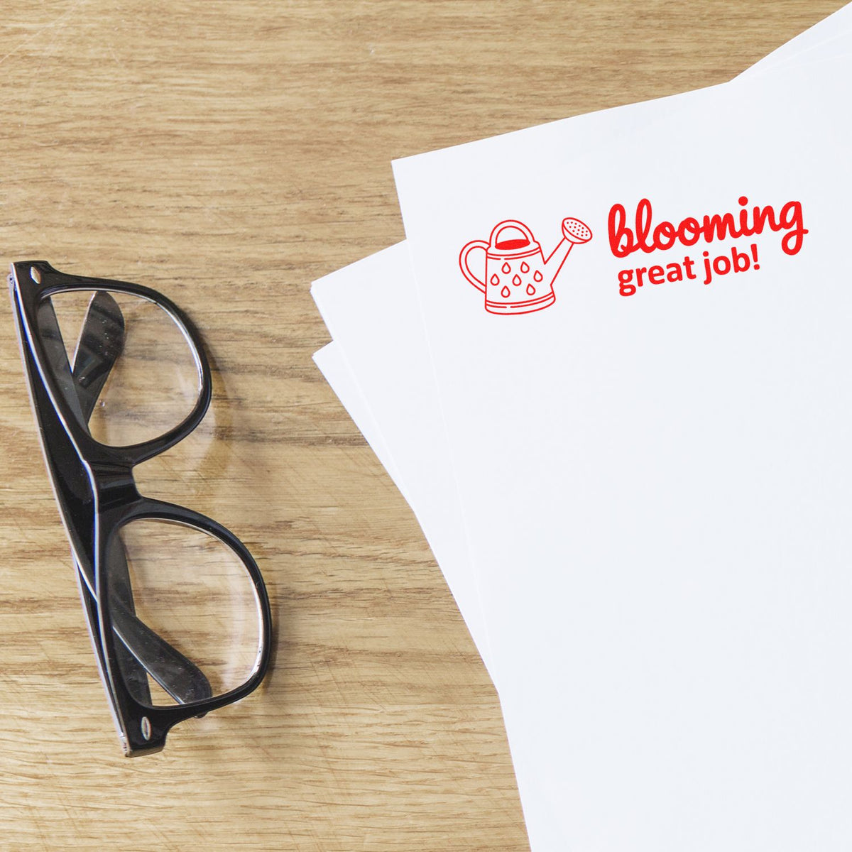 Blooming great job rubber stamp with watering can design on paper, beside eyeglasses on a wooden desk.