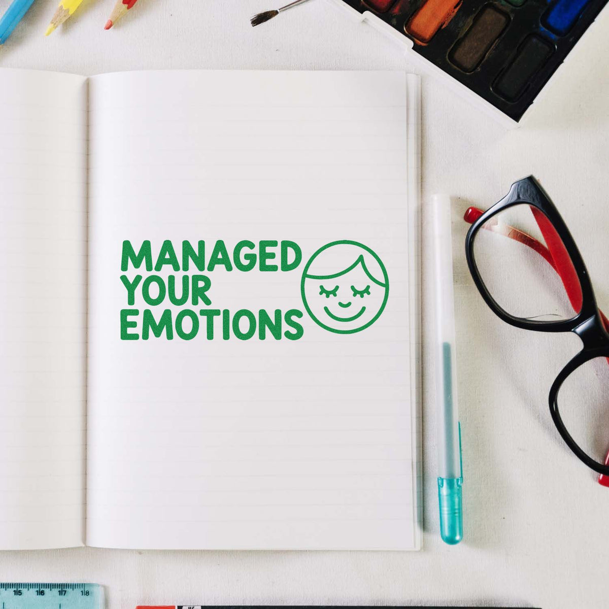 Green rubber stamp saying Managed Your Emotions in a notebook, with smiley face icon and art supplies.