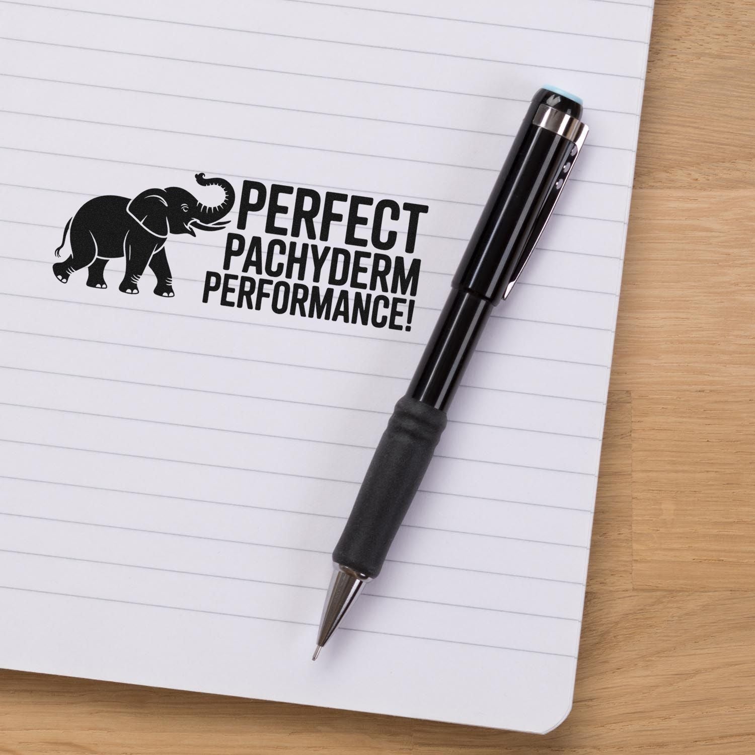 Custom rubber stamp with elephant graphic and Perfect Pachyderm Performance! slogan on a lined notebook beside a pen.