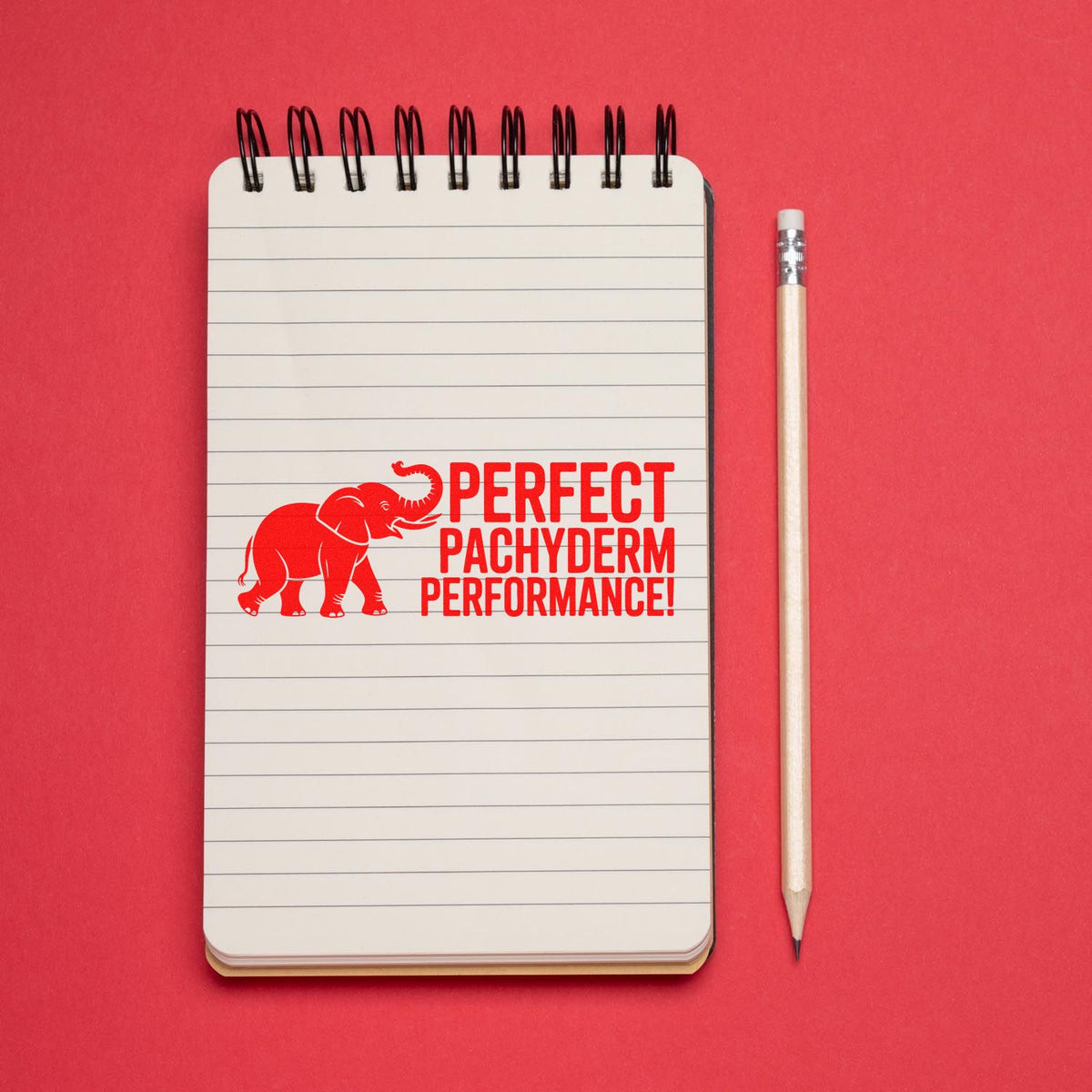 Red elephant rubber stamp slogan Perfect Pachyderm Performance! on a lined notebook with pencil.