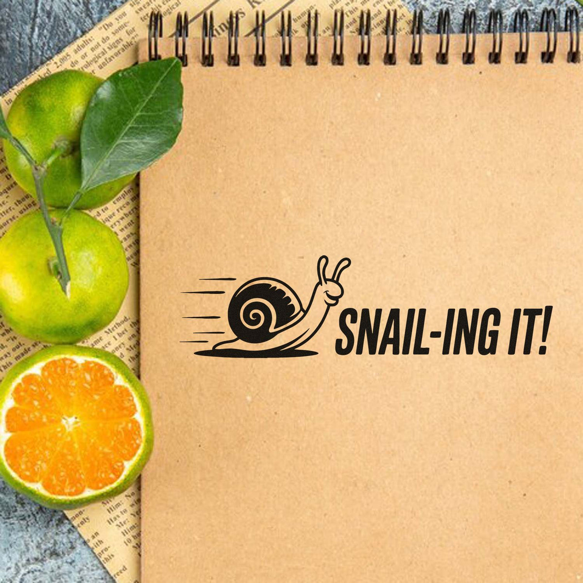 Cute snail rubber stamp design reading Snail-ing It! on a kraft notebook, ideal for journaling, planners, and craft projects.